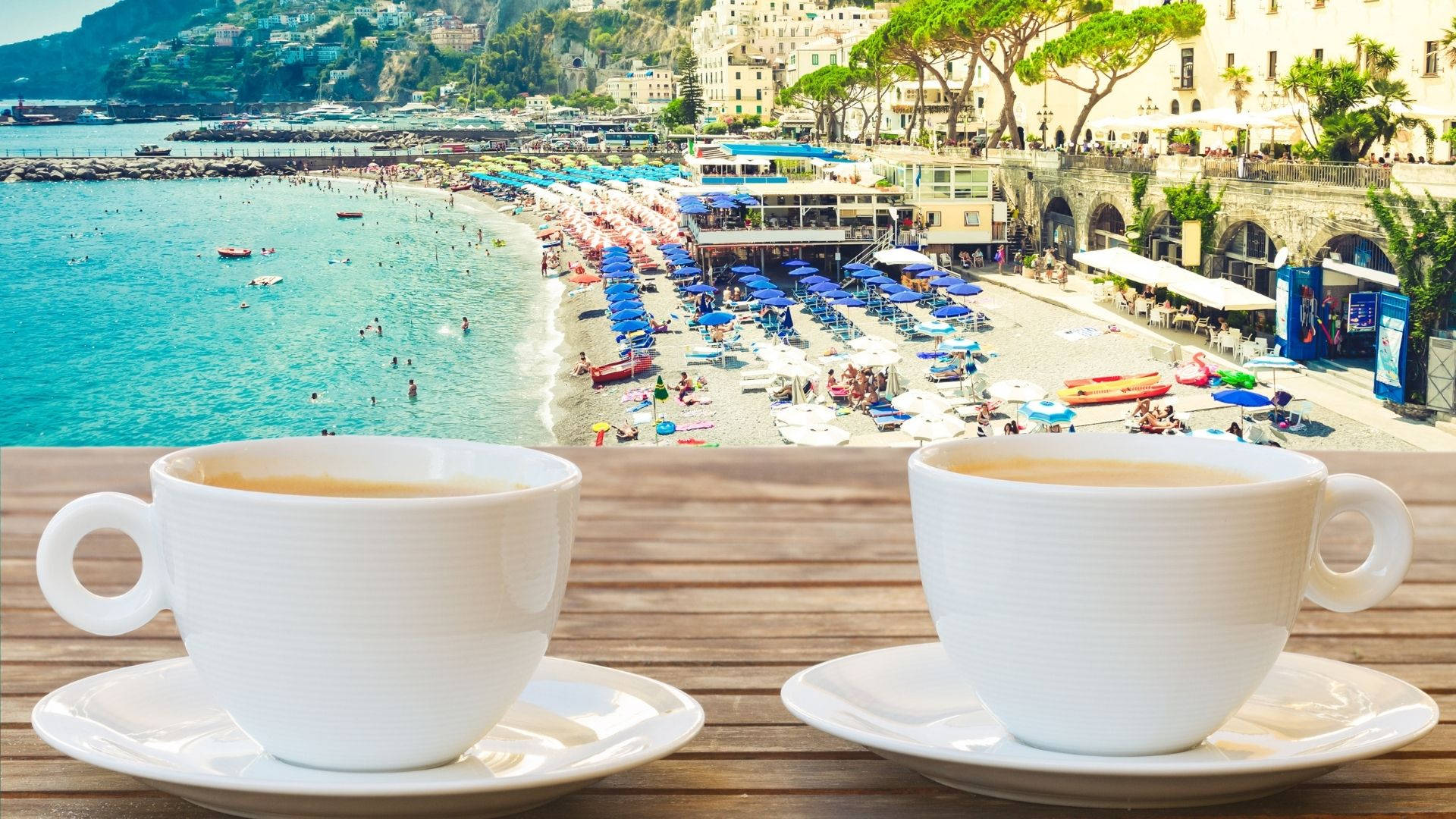 Amalfi Coast 桌布 壁紙 — 兩個白色咖啡杯放在木桌上，俯瞰著充滿活力的海灘場景，有碧綠的海水和色彩繽紛的遮陽傘。
