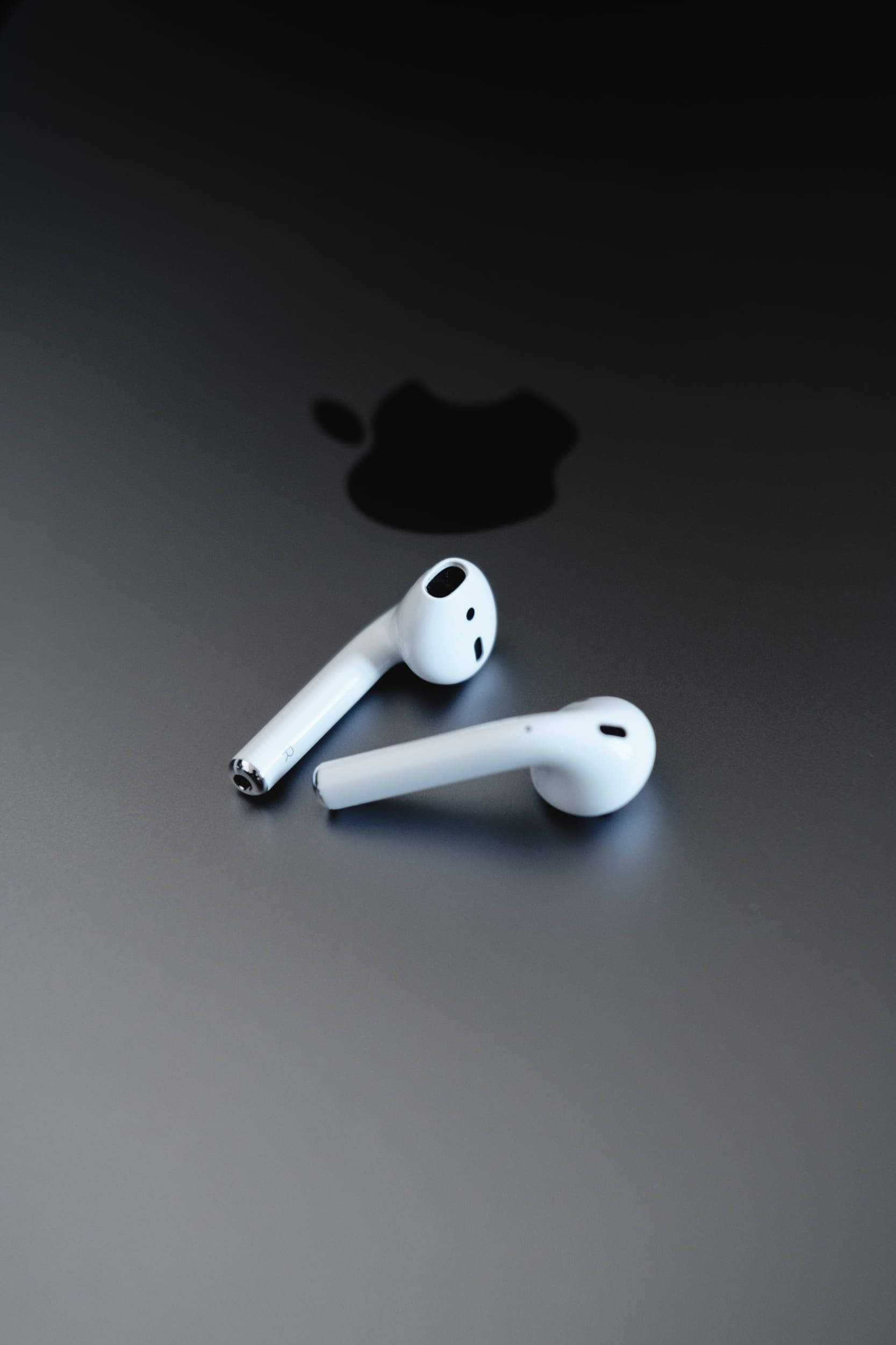 Airpods 桌布 壁紙 — 兩個白色無線耳機放在深色表面上，背後有模糊的蘋果標誌陰影。