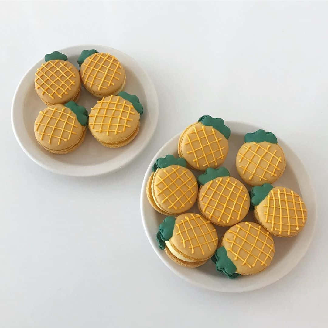 Pineapple 桌布 壁紙 — 兩個白色盤子盛裝著金色鳳梨形馬卡龍，頂部綠色，背景為純白色。