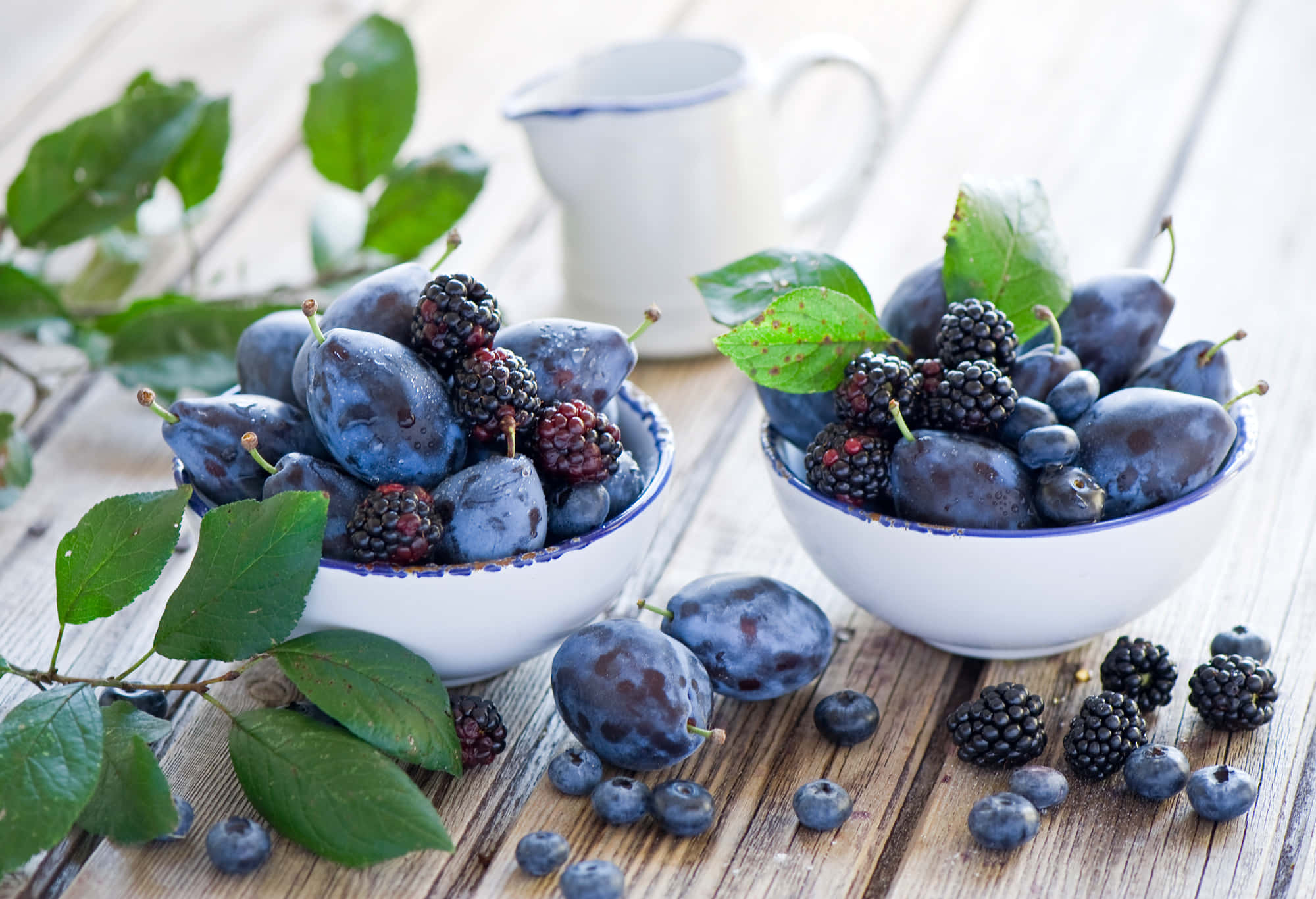 Fruits 桌布 壁紙 — 兩個白色碗裝滿了李子和黑莓，放在一張風化的木桌上，伴隨著新鮮的綠葉。