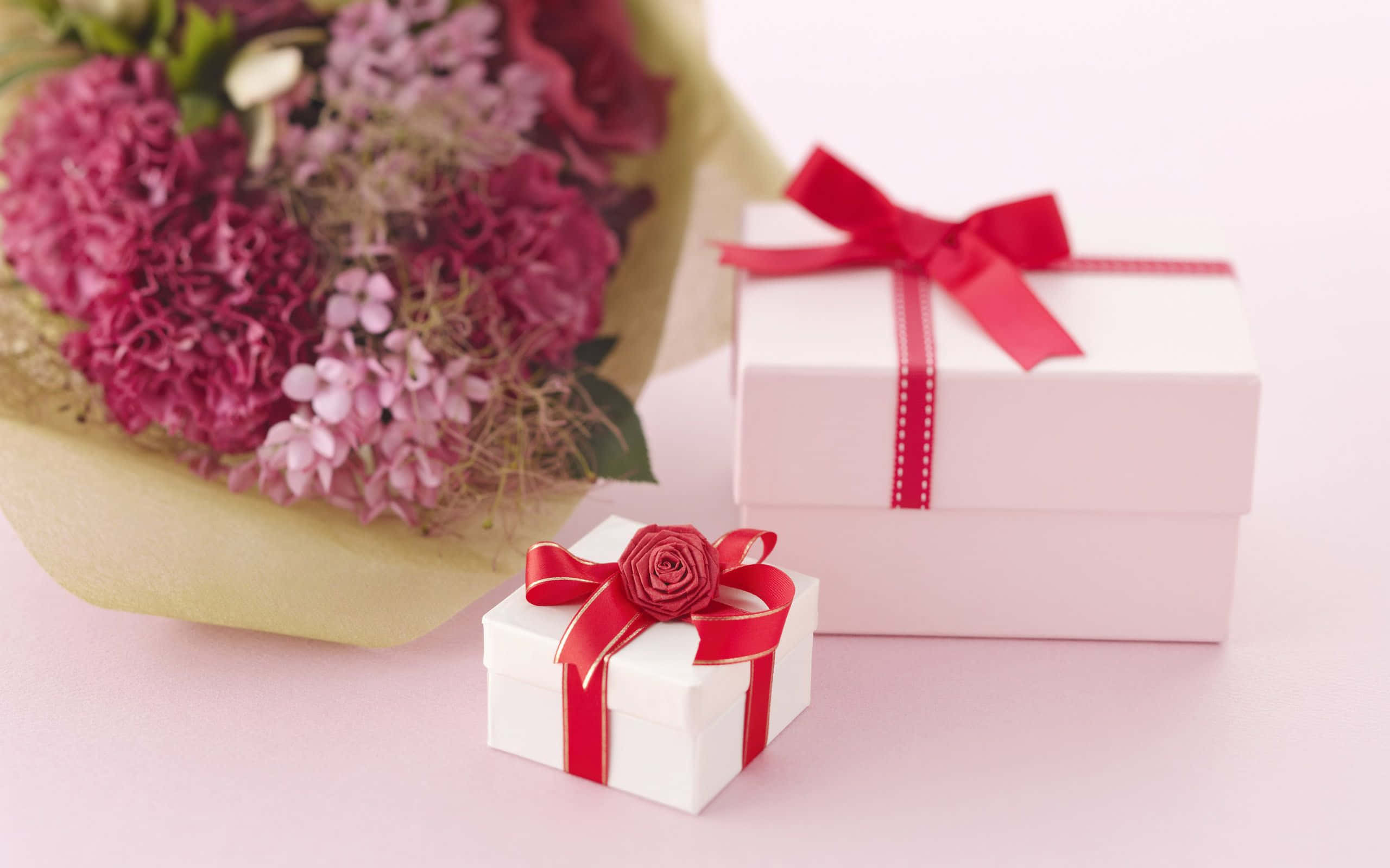 Gift 桌布 壁紙 — 兩個白色禮物盒，帶紅色絲帶，放在粉紅色和紫色繡球花花束旁邊，置於柔軟的粉紅色表面上。