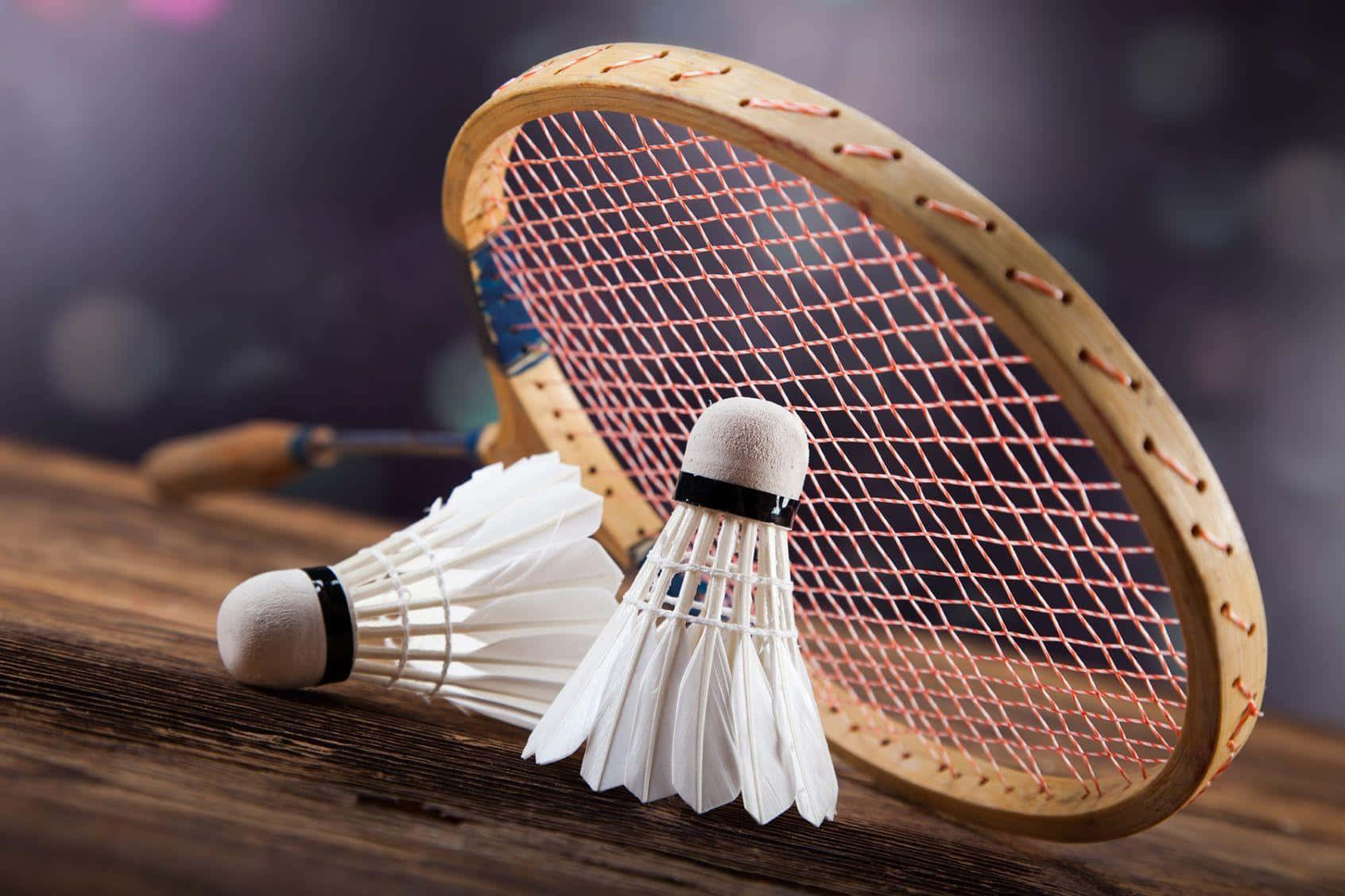 Best Badminton 桌布 壁紙 — 兩個白色羽毛毽子放在一個帶有粉紅色線的木製羽毛球拍旁邊，置於質樸的木質表面上。