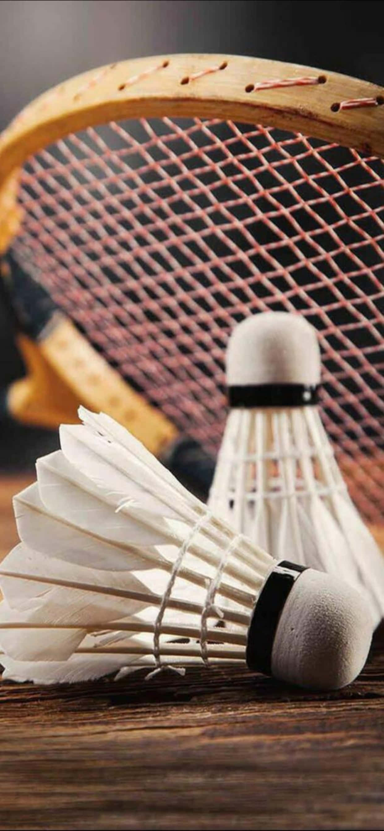 iPhone Xs Max Badminton 桌布 壁紙 — 兩個白色羽毛球放在木質表面上，背景中有一個模糊的羽毛球拍。