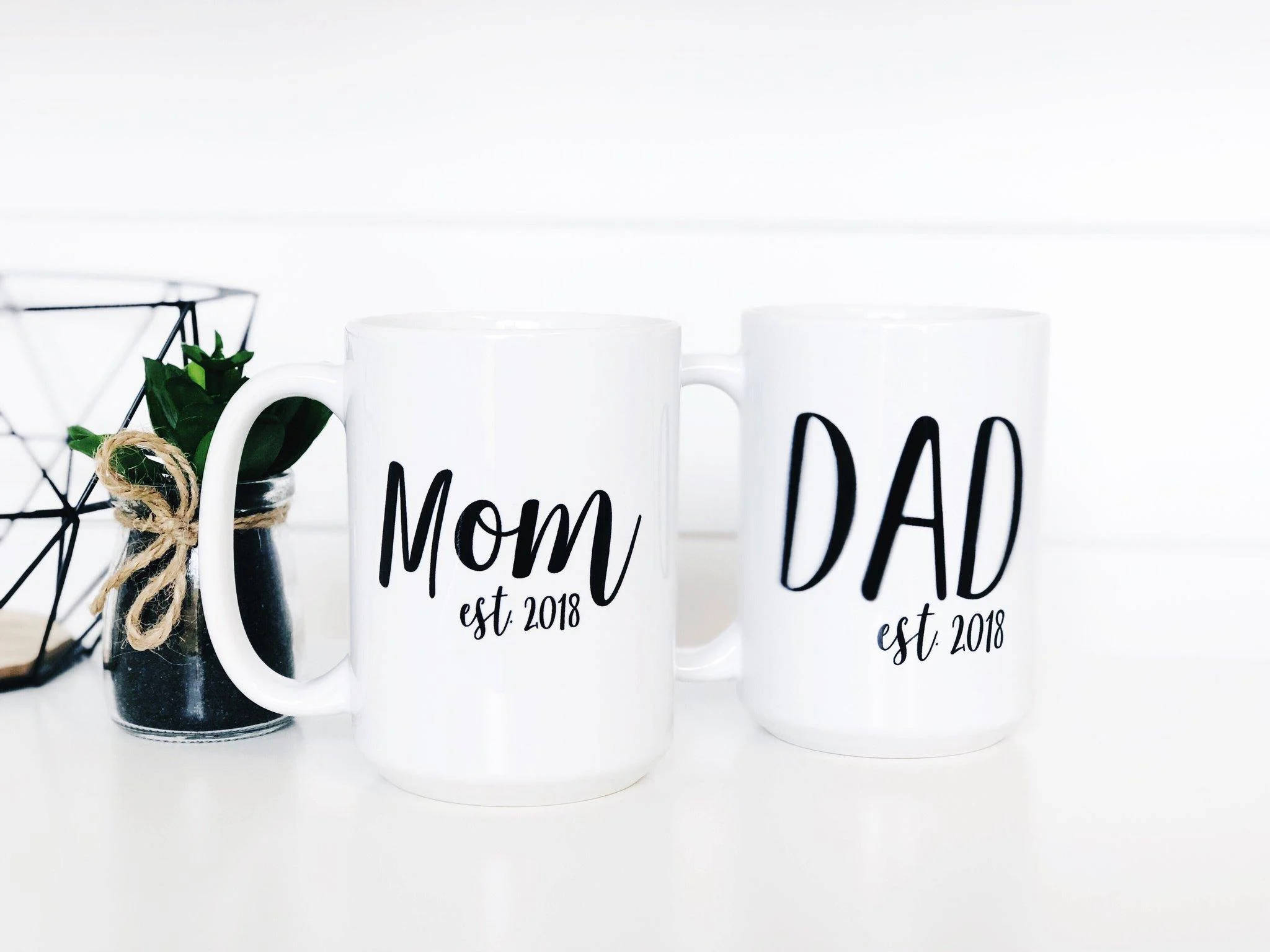 Mom 桌布 壁紙 — 兩個白色馬克杯，上面寫著 Mom and Dad est 2018，旁邊放著一個幾何線條鐵籃裡的小盆栽，放在明亮的白色表面上。