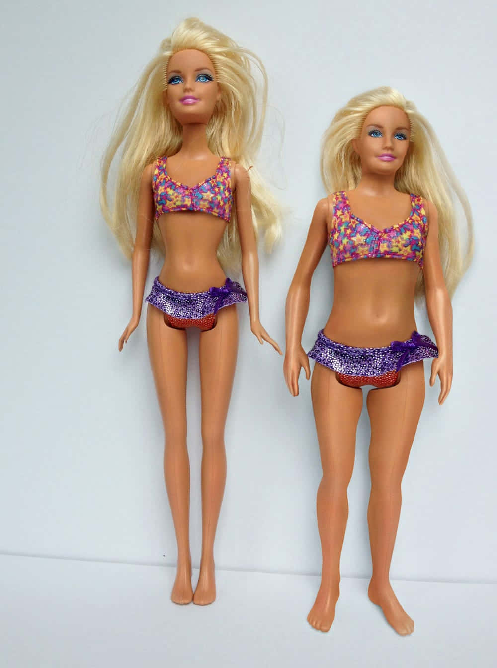 Dolls 桌布 壁紙 — 兩個相同的金髮芭比娃娃穿著相配的彩色比基尼，並排站在純白色背景前。