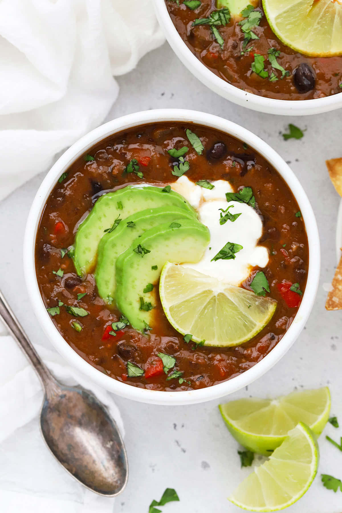 Black Bean Soup 桌布 壁紙 — 兩個碗裝著濃郁的深色辣椒，上面放著酪梨青檸醬和香菜，放在淺灰色表面上。
