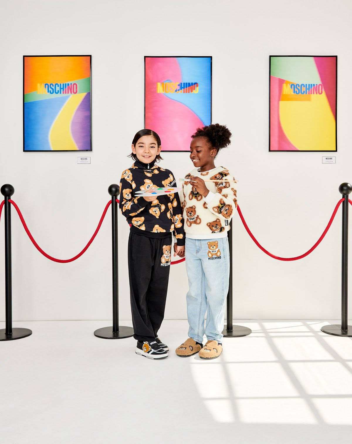 moschino 桌布 壁紙 — 兩個穿著 Moschino 泰迪熊毛衣的孩子在畫廊裡微笑，畫廊裡有色彩繽紛的抽象藝術和紅色天鵝絨繩索。