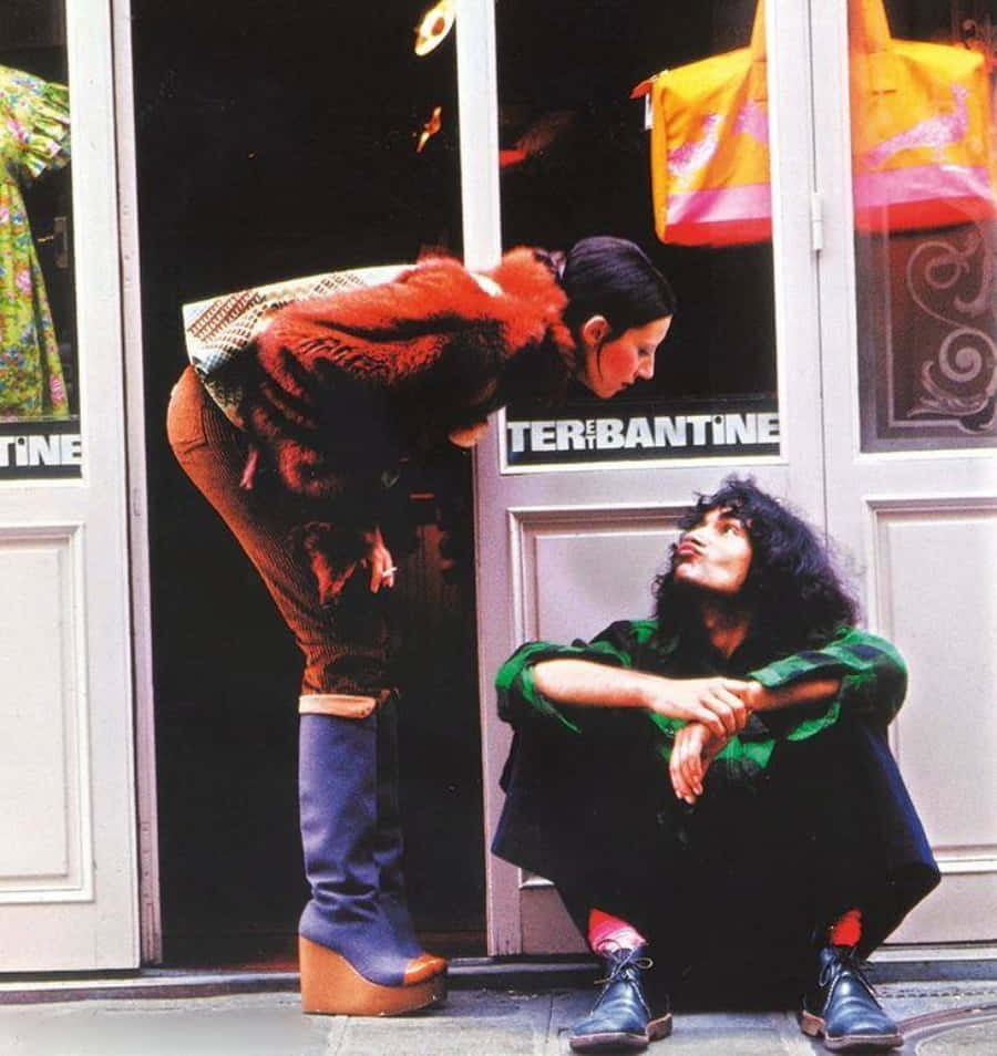 70年代動圖 桌布 壁紙 — 兩個穿著1970年代時尚的人在商店門附近互動，一人蹲下，另一人彎腰穿著厚底靴和毛領。