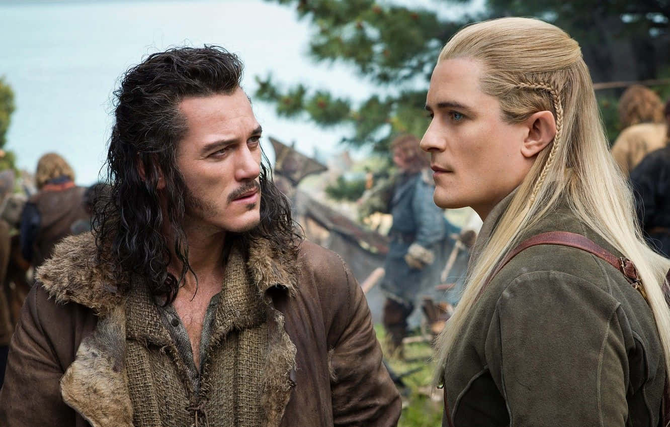 legolas 桌布 壁紙 — 兩個穿著中世紀服裝的男人在戶外營地中面對面，背景有樹木和模糊的人物。
