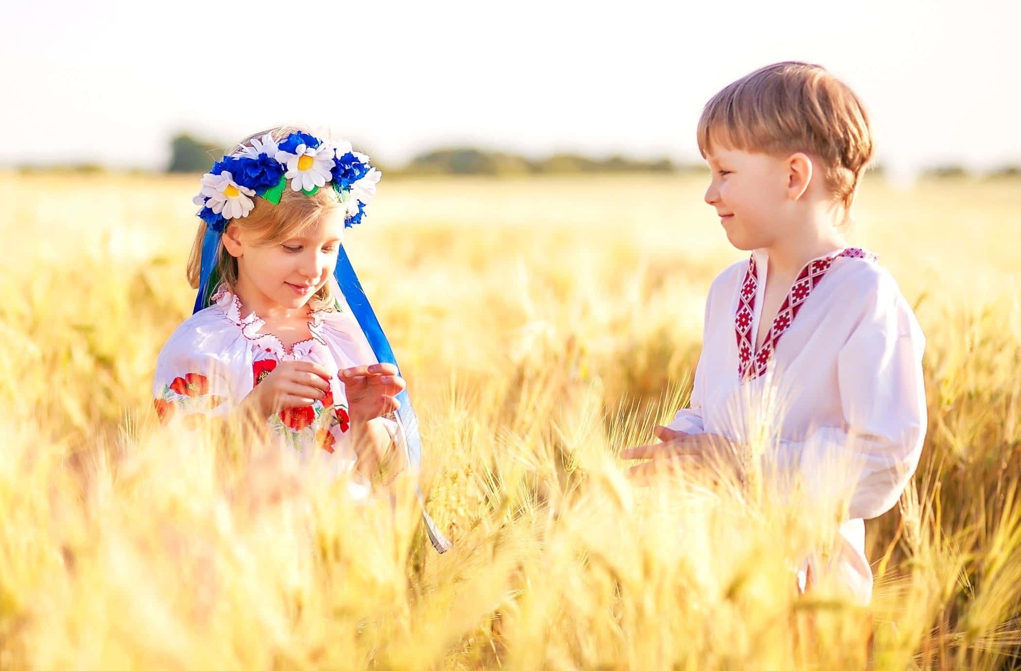 Cute Kids 桌布 壁紙 — 兩個穿著傳統刺繡服裝的孩子站在金色麥田裡，在溫暖的陽光下互相微笑。