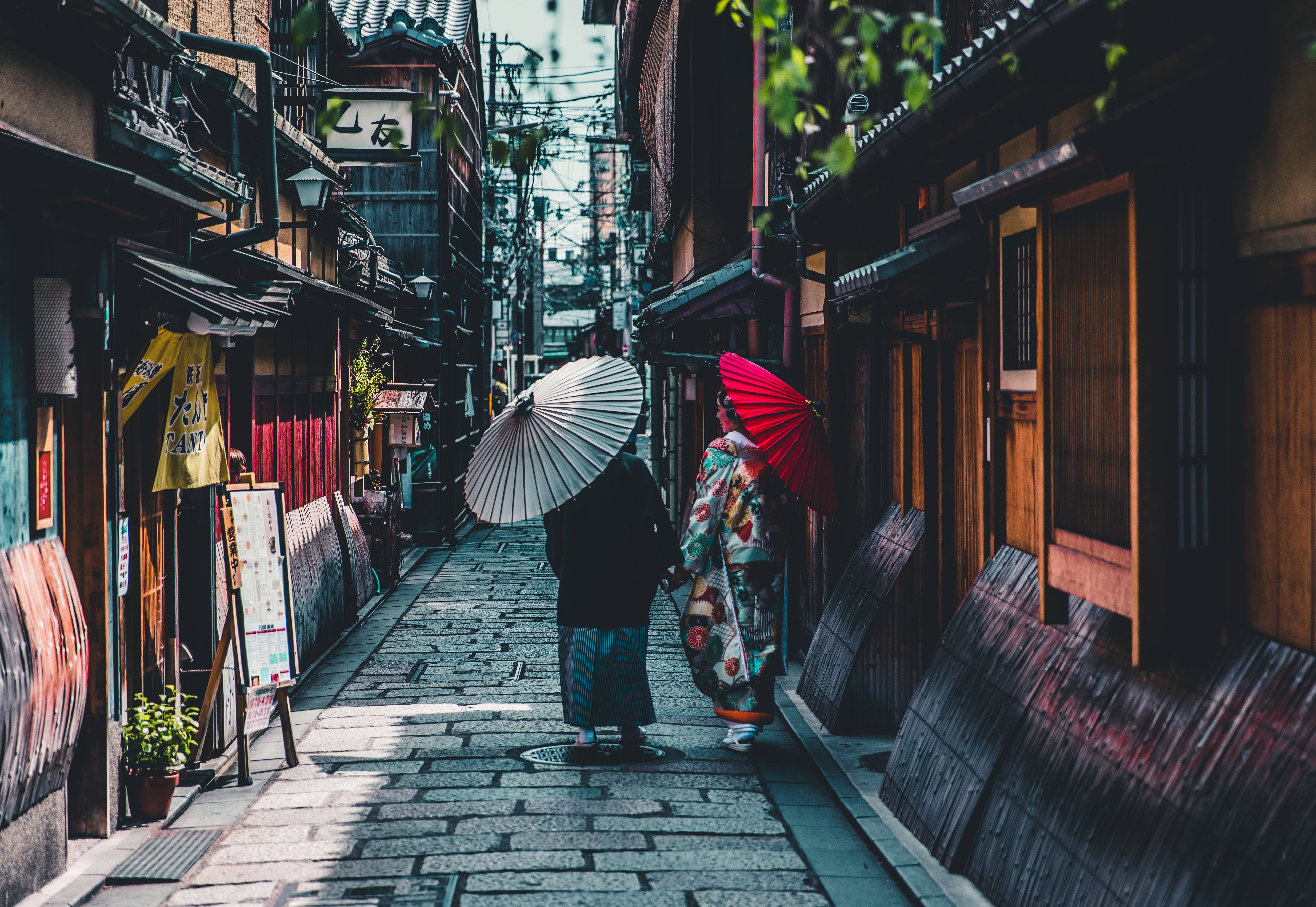日本 桌布 壁紙 — 兩個穿著傳統日本服飾的人物在柔和日光下，沿著一條兩旁有木建築的狹窄鵝卵石小巷行走。