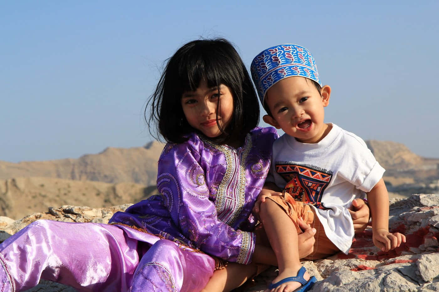 Brother And Sister 桌布 壁紙 — 兩個穿著傳統服飾的孩子坐在岩石地形上，背景是乾旱的山脈和清澈的藍天。