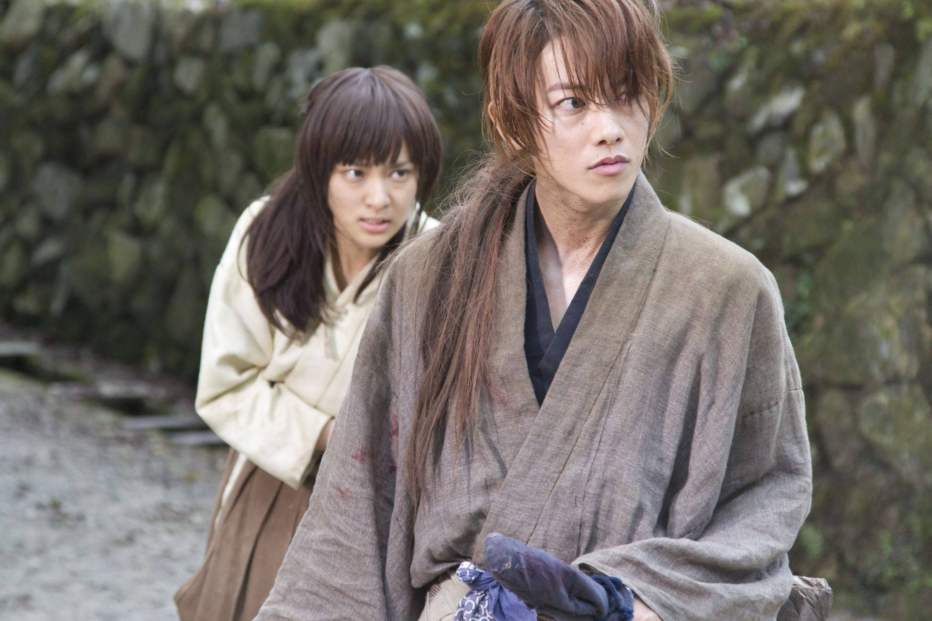 Kenshin 桌布 壁紙 — 兩個穿著傳統長袍的人站在一條石徑上，身後有長滿苔蘚的岩石；其中一人表情嚴肅地向前看，而另一人則擔憂地越過肩膀張望。