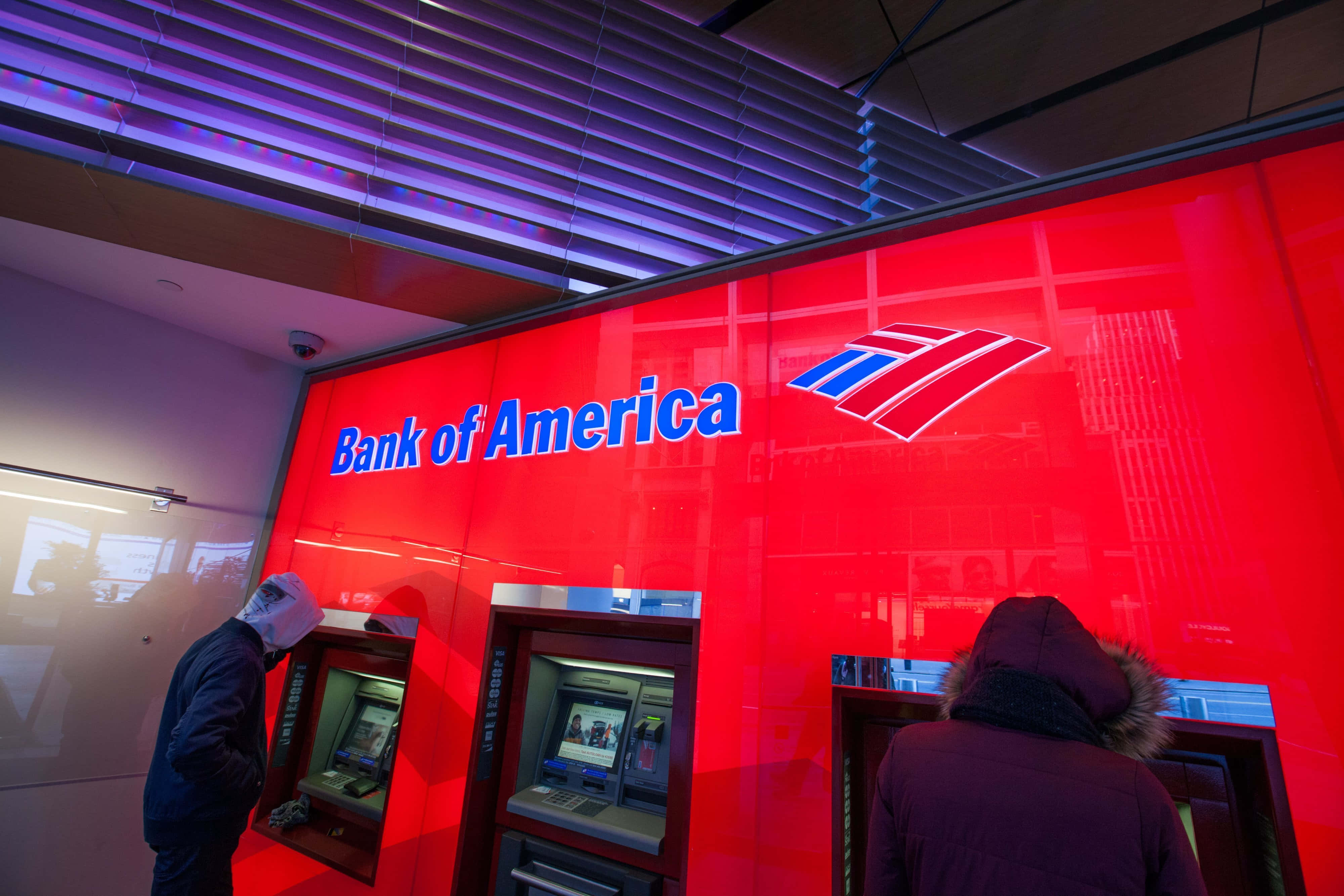 美國銀行 桌布 壁紙 — 兩個穿著冬季外套的人在發光紅色Bank of America標誌和紫色天花板燈光下使用自動櫃員機。