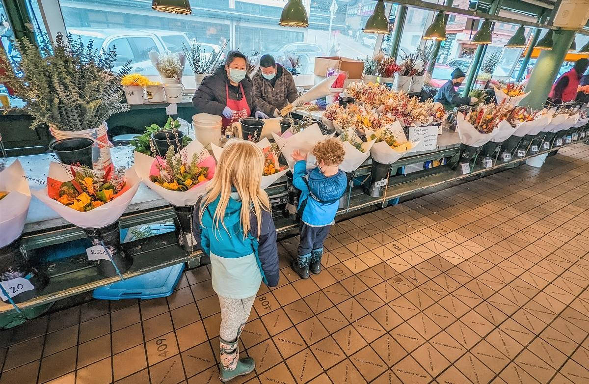 Pike Place Market 桌布 壁紙 — 兩個穿著外套的小孩在繁忙的室內市場攤位前瀏覽色彩繽紛的花束，地板鋪有瓷磚，掛著燈飾。