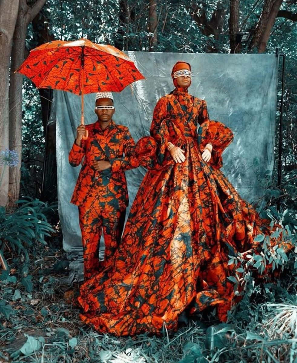 Afrofuturism 桌布 壁紙 — 兩個穿著大膽橙色和黑色圖案服裝的人物站在森林中，其中一人拿著相配的雨傘，背景為灰色。