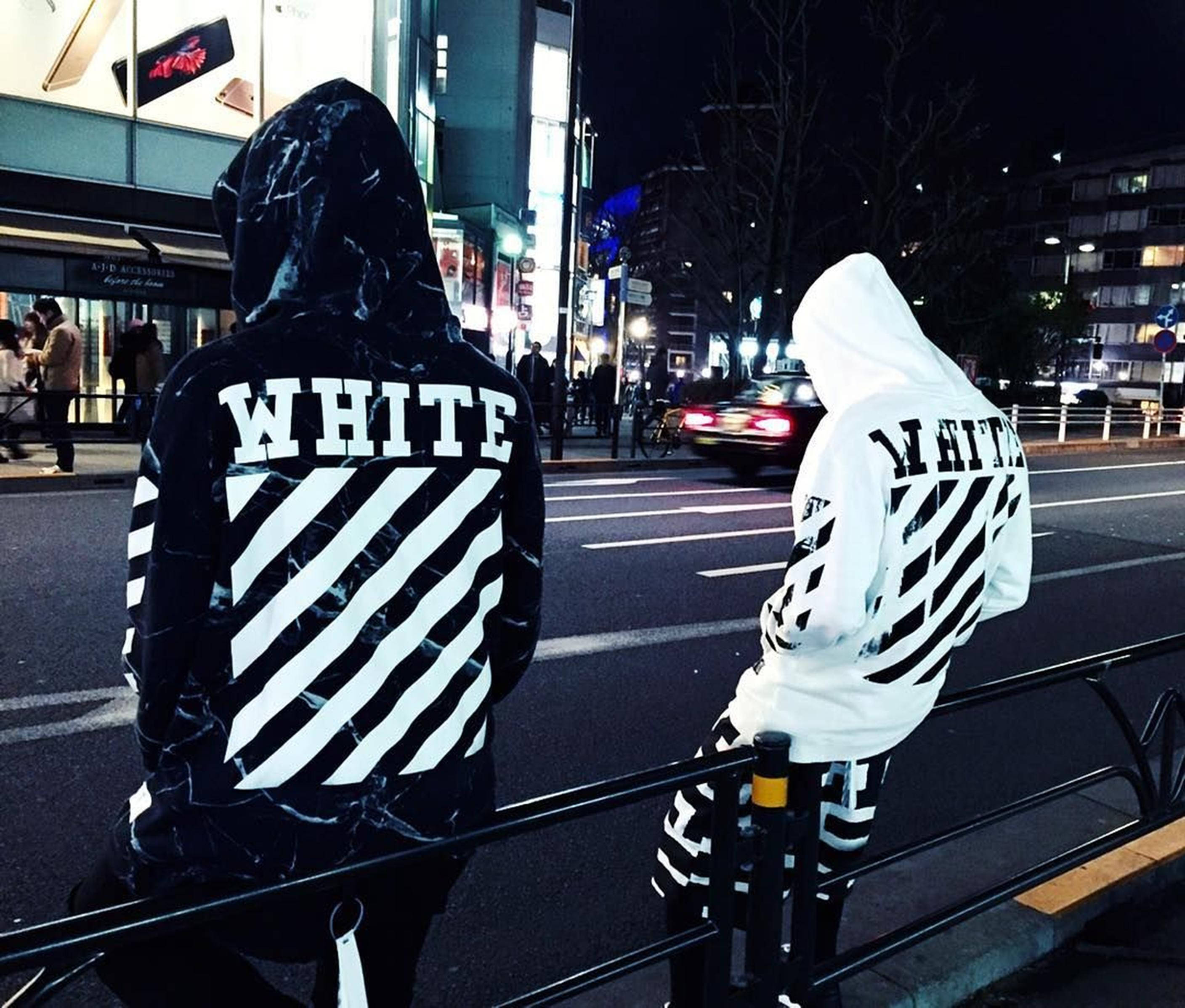 Off White Logo 桌布 壁紙 — 兩個穿著大膽黑白連帽衫的人坐在欄杆上，面向夜晚霓虹燈照亮的城市街道。