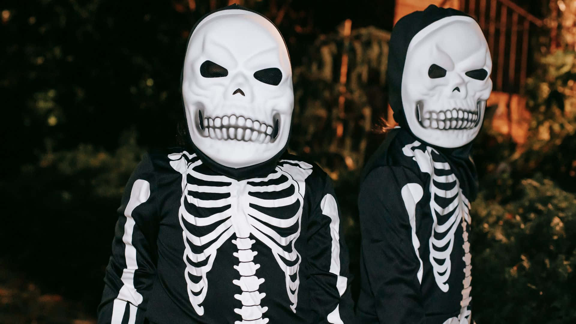 Skeleton Costumes 桌布 壁紙 — 兩個穿著完整骷髏服裝、戴著頭骨面具的孩子在光線昏暗的戶外環境中並肩站立。