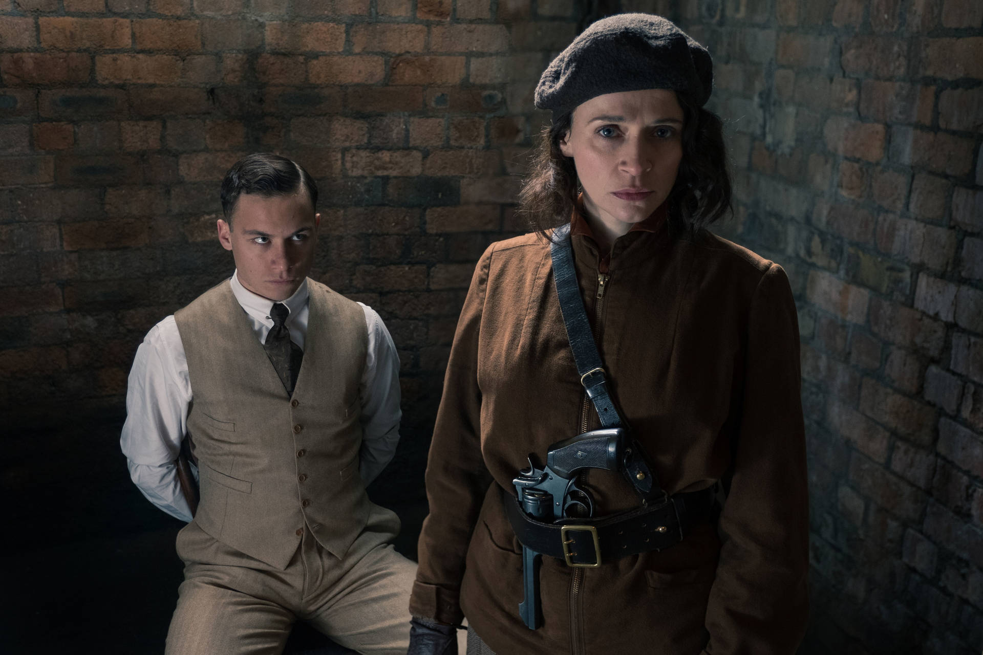 兩個穿著1940年代服裝的人，在昏暗的磚房裡，一個手持手槍。 — Peaky Blinders 壁紙