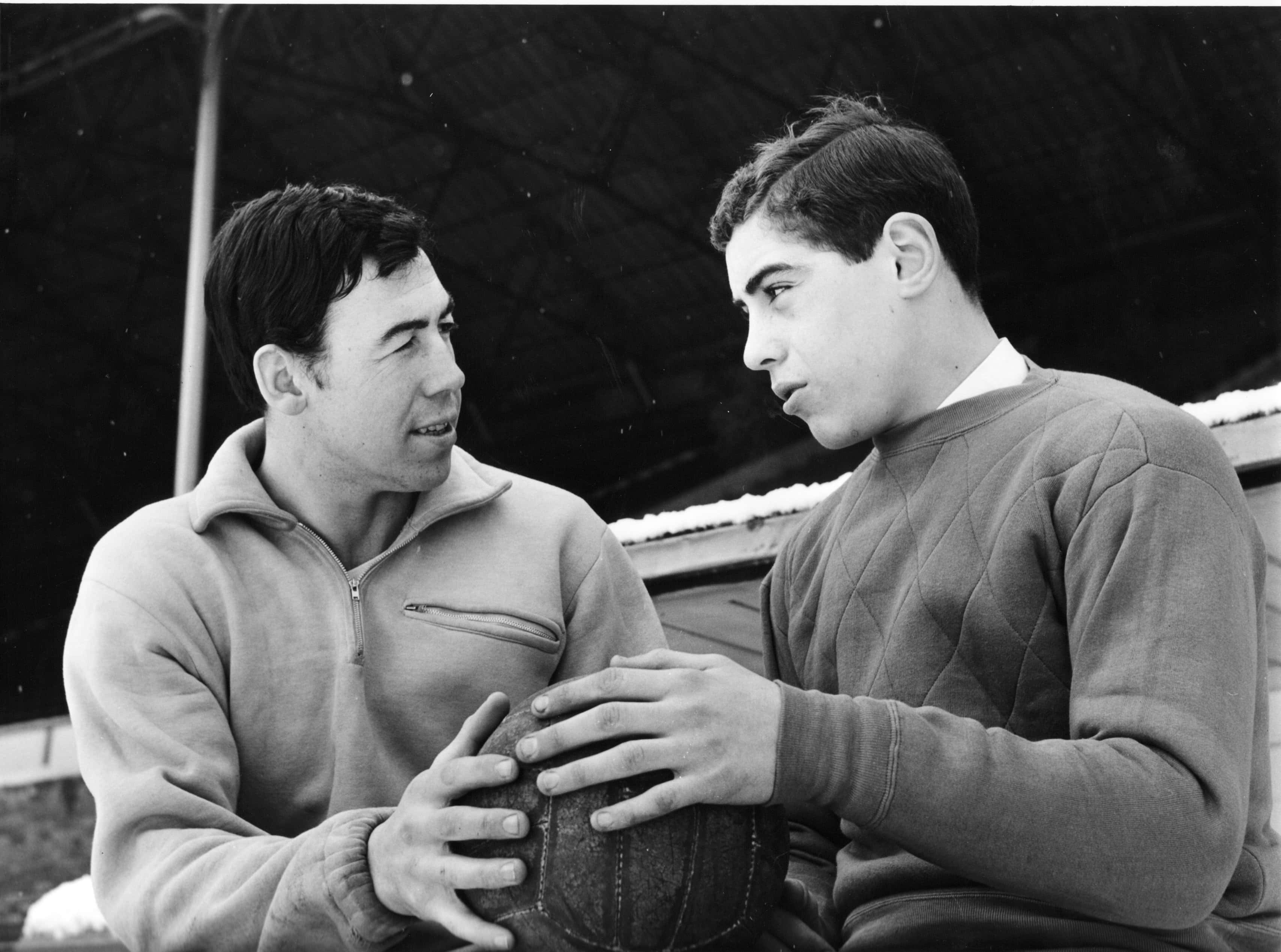 Football Players 桌布 壁紙 — 兩個穿著復古毛衣的男人在一個有蓋結構下拿著足球進行專注的對話。