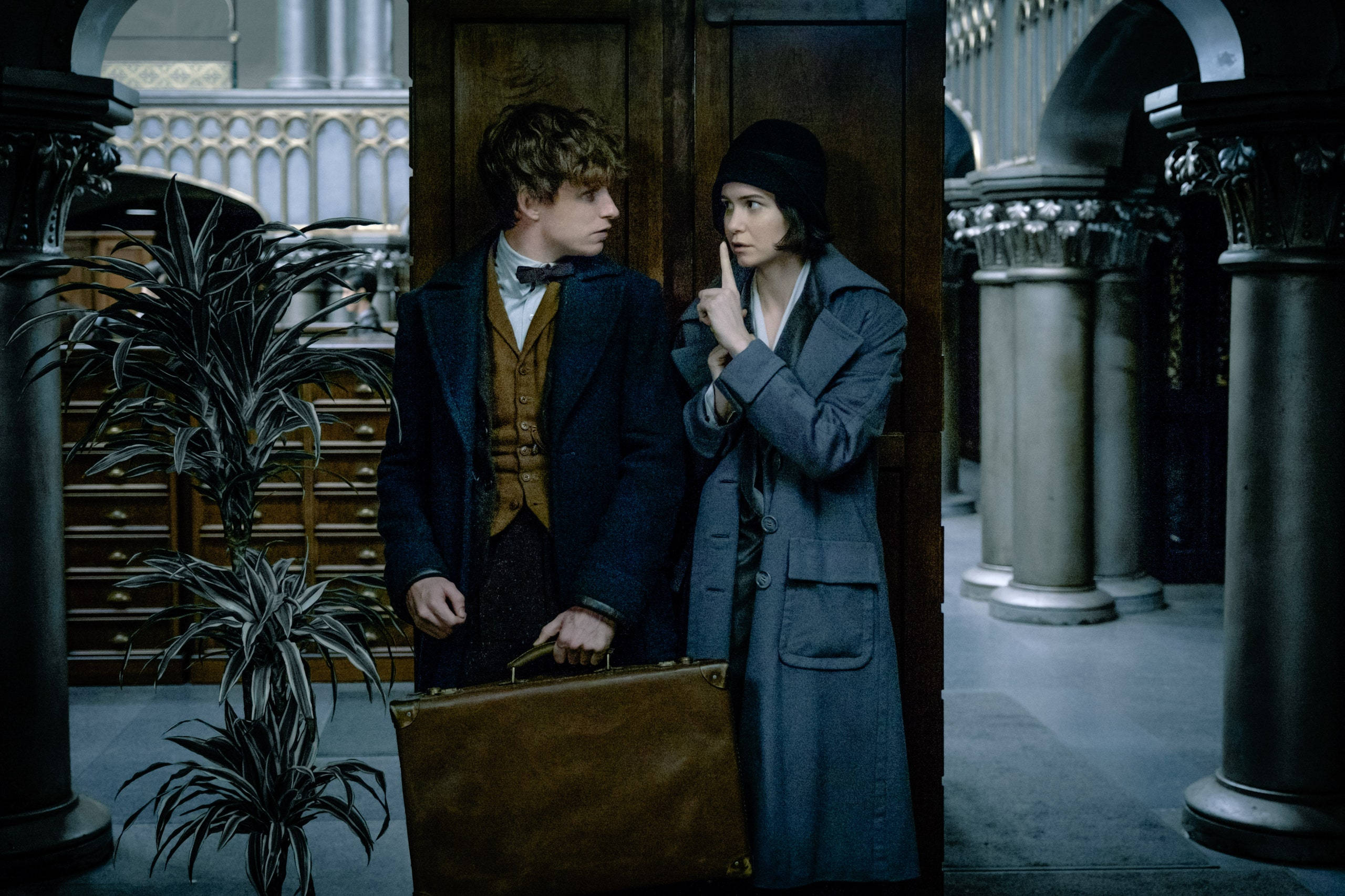 電影動圖 桌布 壁紙 — 兩個穿著時代服裝的人物站在宏偉的大廳中，一個拿著手提箱，另一個無聲地將手指放在嘴唇上做手勢。