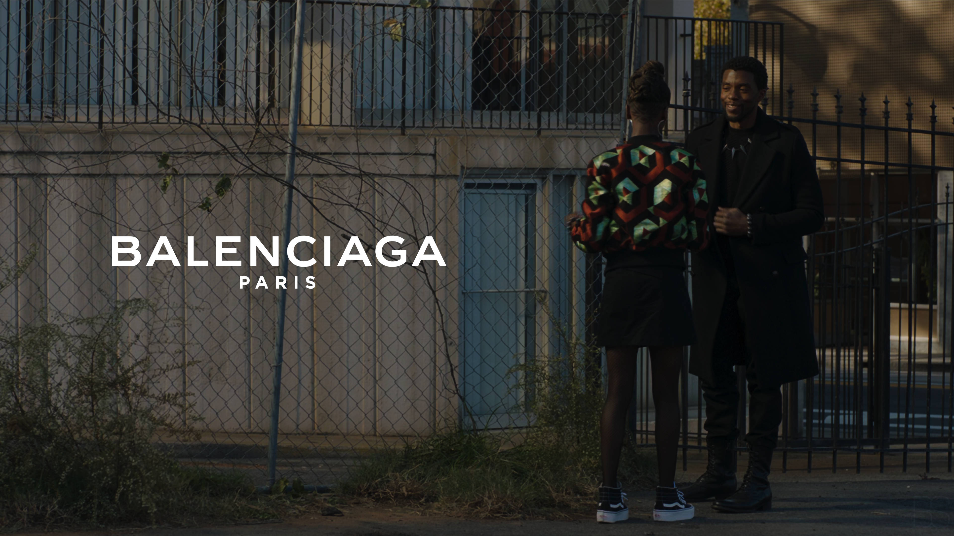 Balenciaga 桌布 壁紙 — 兩個穿著時尚的人站在鐵絲網圍欄附近，帶有「Balenciaga Paris」文字疊加在粗糙的城市環境中。