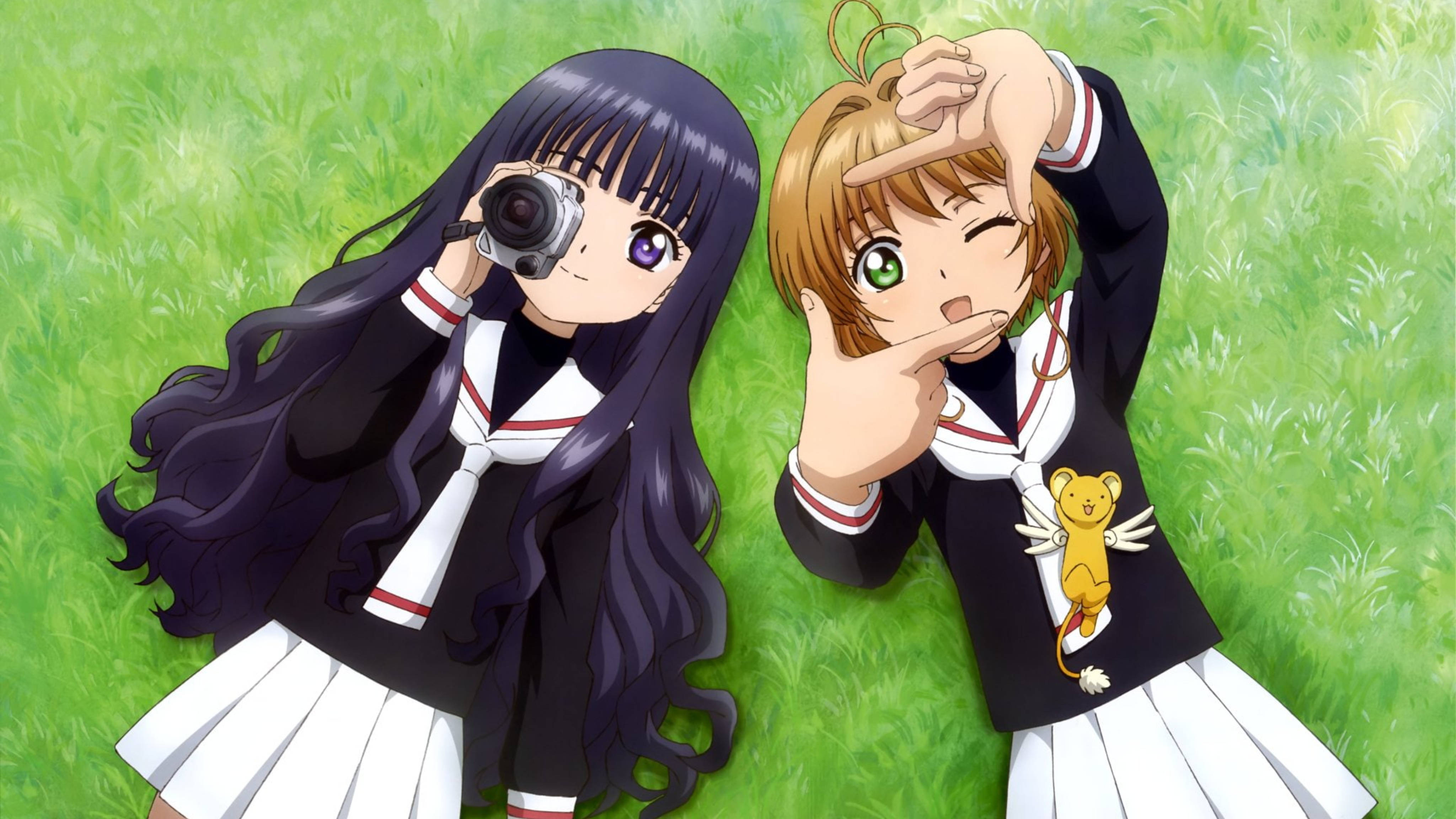 Cardcaptor 桌布 壁紙 — 兩個穿著校服的動漫女孩在充滿活力的綠色草地上擺出俏皮的姿勢，一個拿著相機，另一個用手做出心形。