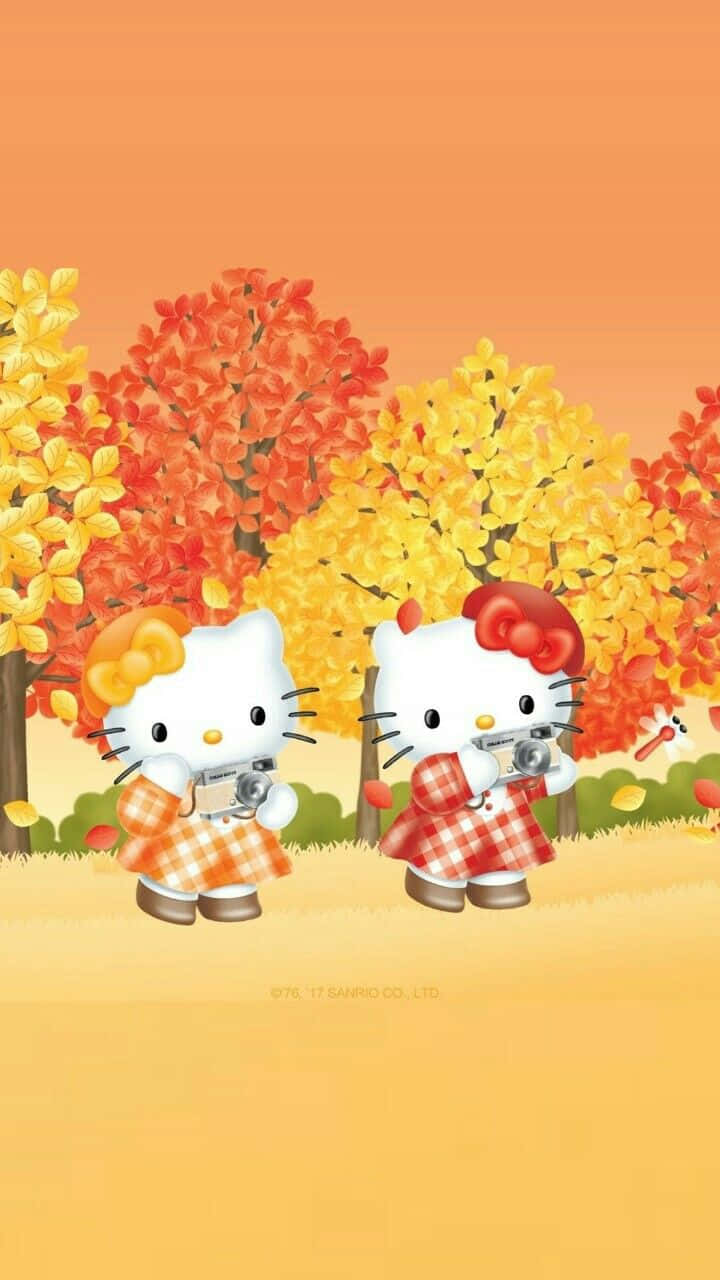 Hello Kitty Thanksgiving 桌布 壁紙 — 兩個穿著格紋服裝的Hello Kitty角色在溫暖橙色天空下，於鮮豔的秋樹前手持相機。