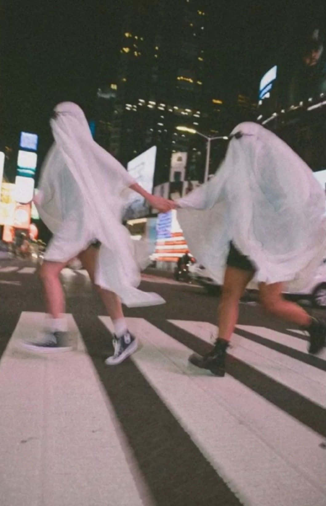 Best Friend Aesthetic 桌布 壁紙 — 兩個穿著白色鬼魂服裝的人手牽手，在夜晚穿過一條霓虹燈照亮的城市街道。