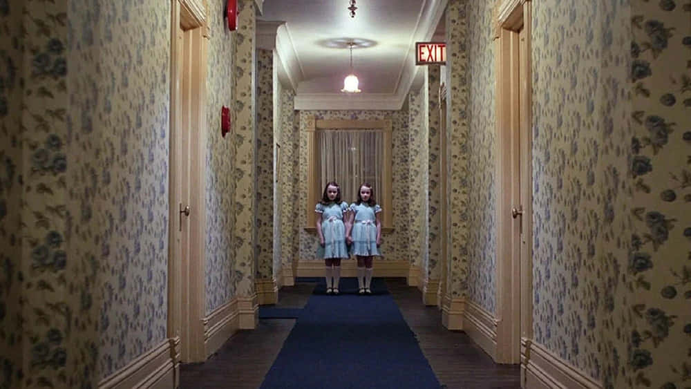 Haunted Hotels 桌布 壁紙 — 兩個穿著相同連衣裙的年輕女孩，詭異地站在一條長長的復古走廊盡頭，走廊上有花卉壁紙和上方發光的出口標誌。
