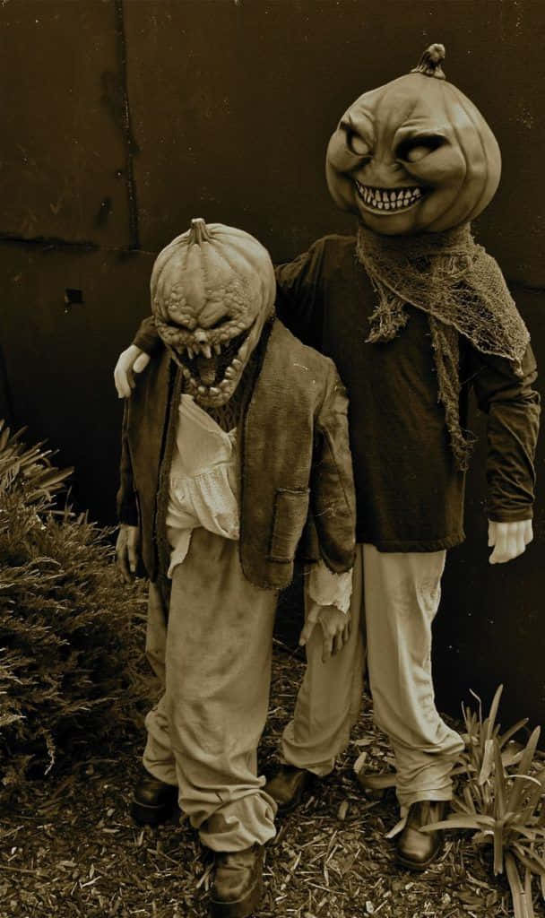 Creepy Halloween 桌布 壁紙 — 兩個穿著破爛衣服、頭戴南瓜頭的詭異人物，在深褐色調的暗牆前威嚇地站立。