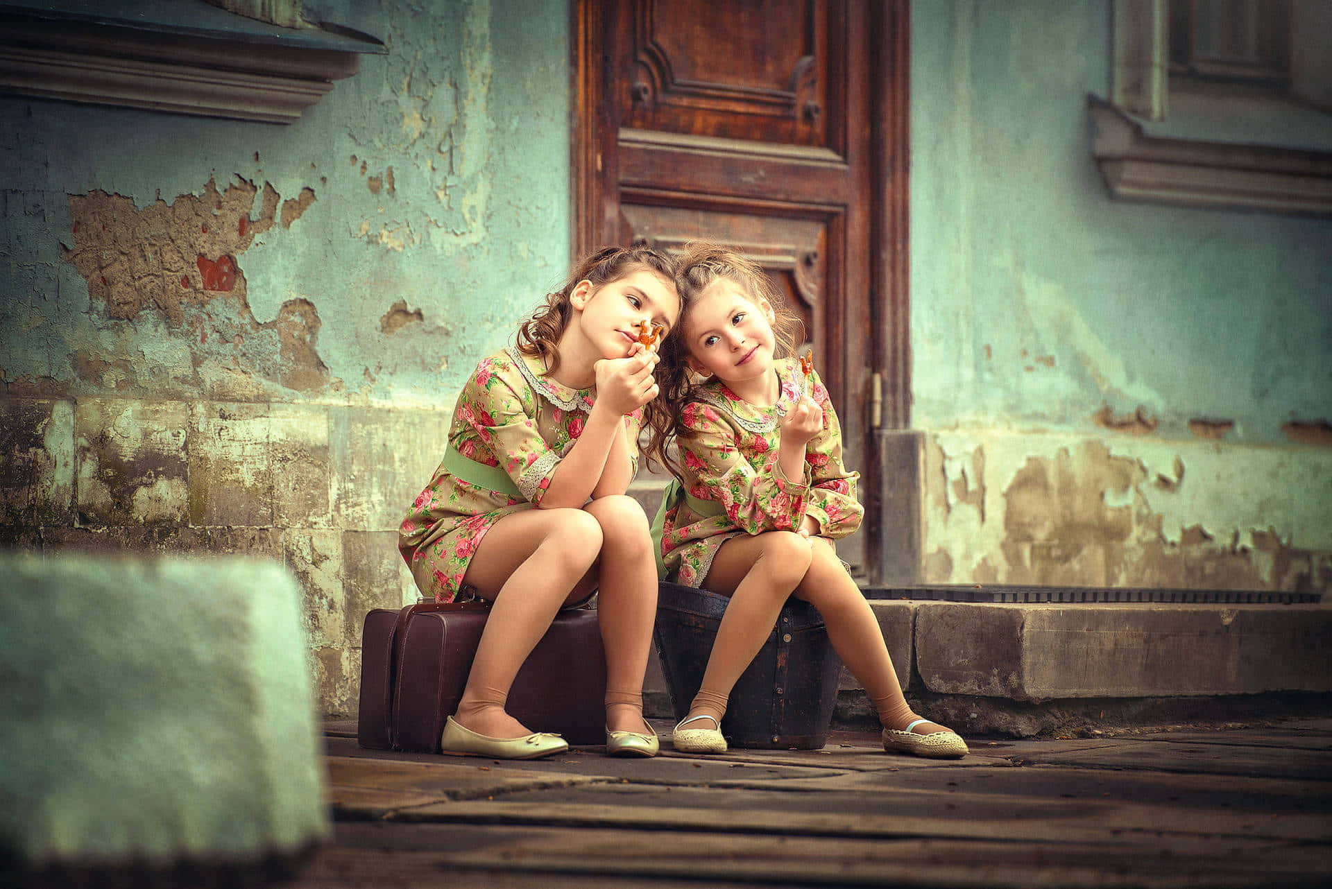 sisters 桌布 壁紙 — 兩個穿著碎花裙的小女孩坐在行李箱上，背景是風化的青綠色牆壁，分享著甜蜜的時刻。