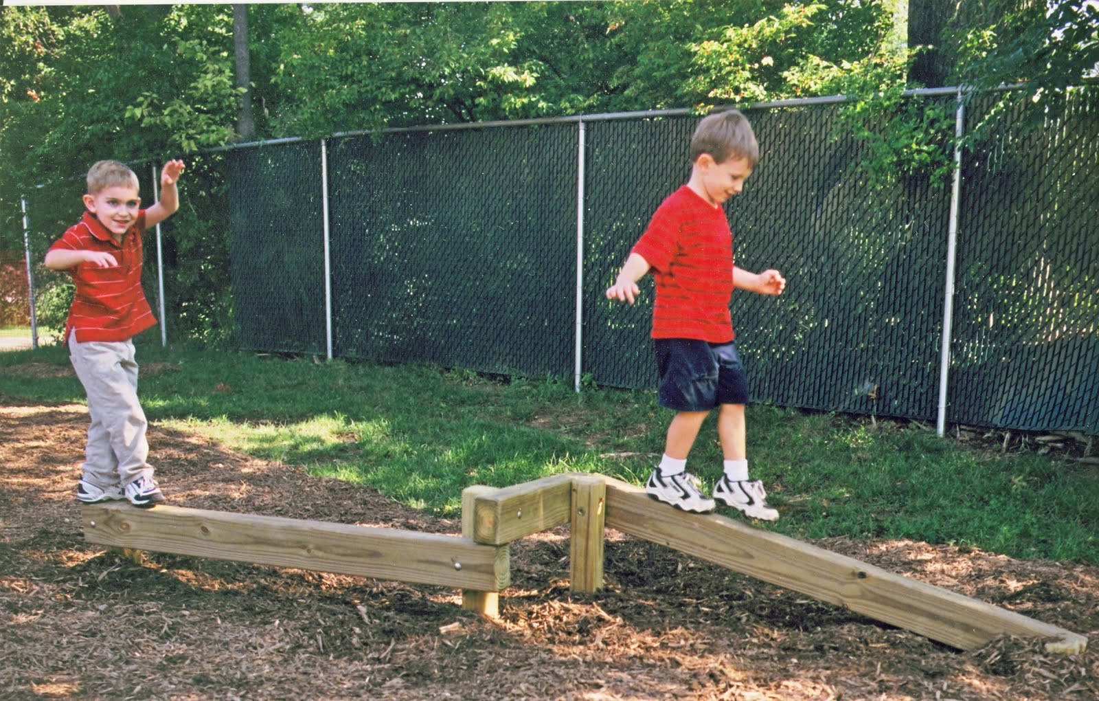 Playground 桌布 壁紙 — 兩個穿著紅色襯衫的男孩在陽光明媚的後院遊樂場的木樑上平衡玩耍。