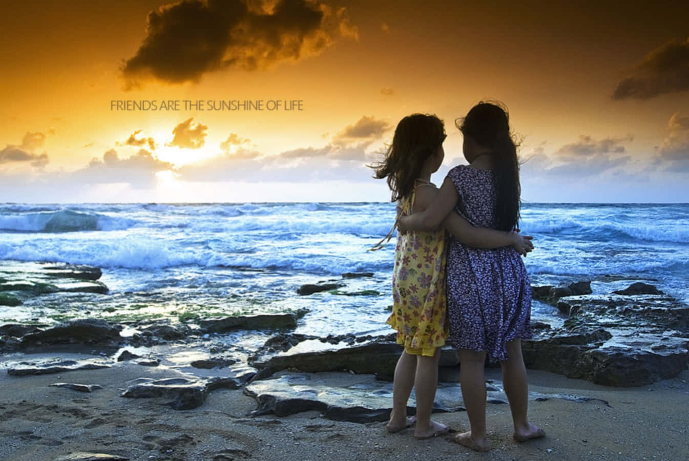 Friends 桌布 壁紙 — 兩個穿著花卉裙的女孩在岩石海灘上擁抱，金色夕陽照耀著海浪。