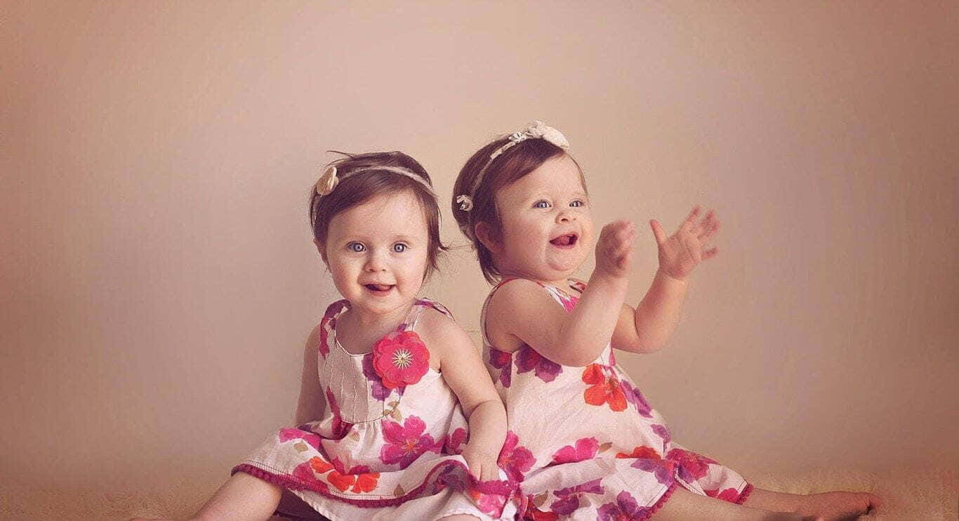Cute Babies 桌布 壁紙 — 兩個穿著花卉連衣裙的快樂幼兒背對背坐在柔軟的米色背景前，揮手並燦爛地微笑。