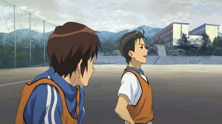 Haruhi Taniguchi 桌布 壁紙 — 兩個穿著足球制服的動畫男孩在學校球場上交談，背景有山脈和建築物。