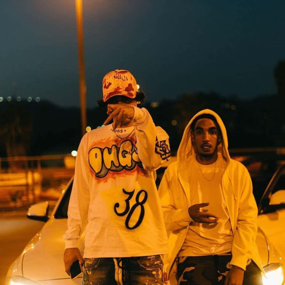 Shoreline Mafia 桌布 壁紙 — 兩個穿著連帽衫和帽子的年輕男子在夜晚溫暖的路燈下自信地站在車旁擺姿勢。