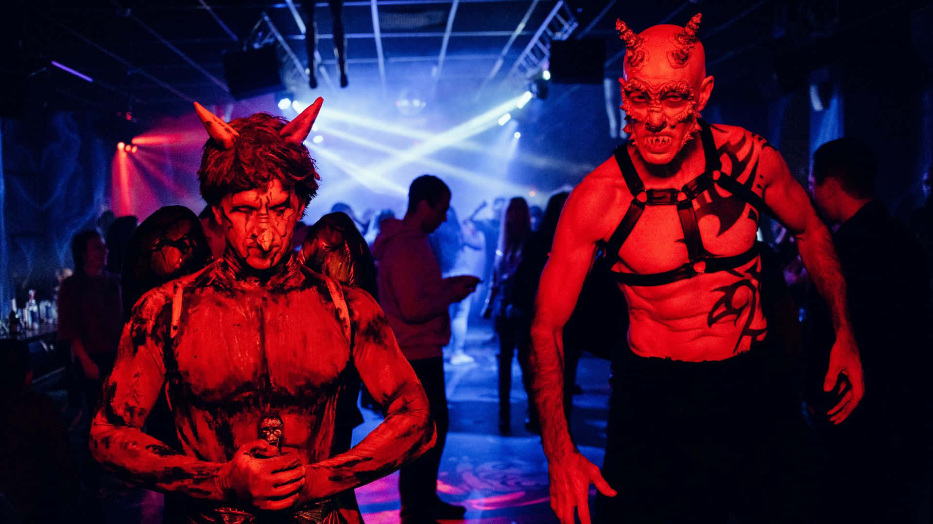 Horror Costumes 桌布 壁紙 — 兩個穿著魔鬼服裝、臉上塗著紅色顏料、頭上有角的男人站在燈光昏暗的俱樂部裡，在閃爍的藍色和紅色燈光下。
