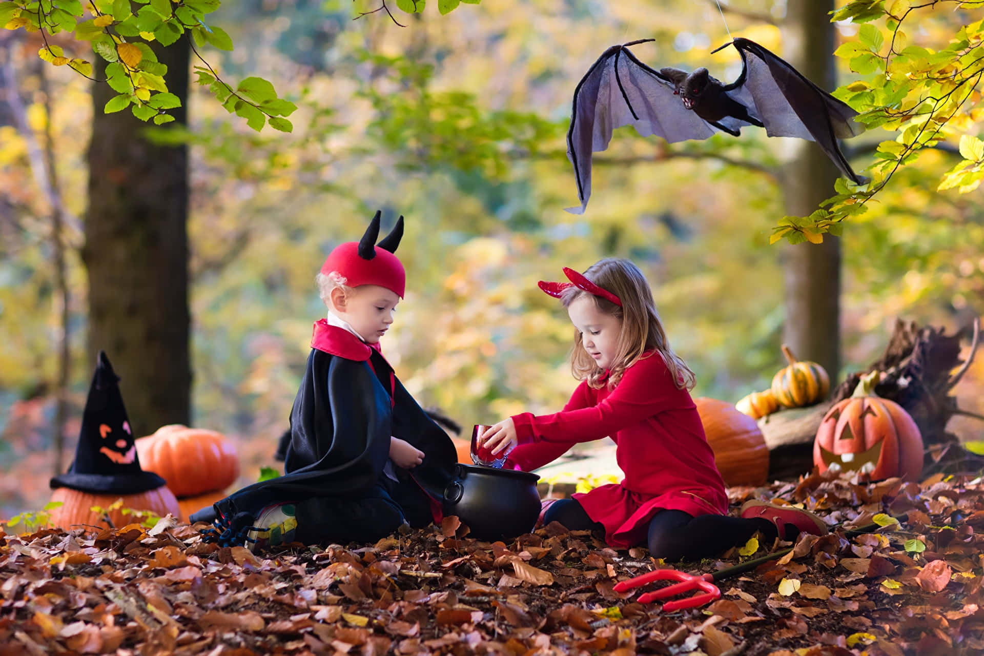 Halloween Costume 桌布 壁紙 — 兩個穿著魔鬼服裝的小孩在蝙蝠下，於秋葉和南瓜中攪拌大鍋中的藥水。