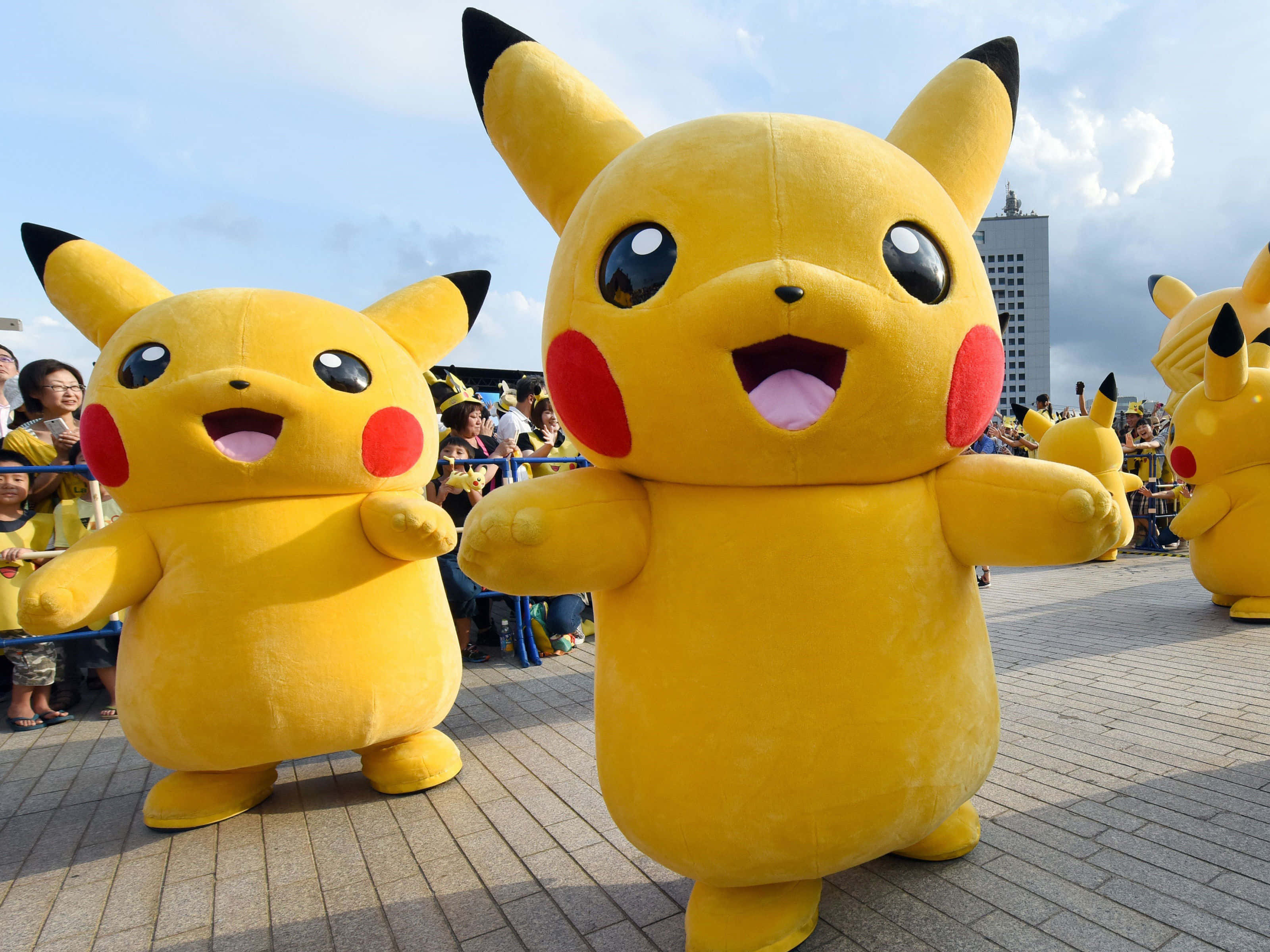 Pokemon Toys 桌布 壁紙 — 兩個穿著黃色皮卡丘服裝的開心角色，在鋪砌的廣場上微笑站立，身後有群眾和建築物。