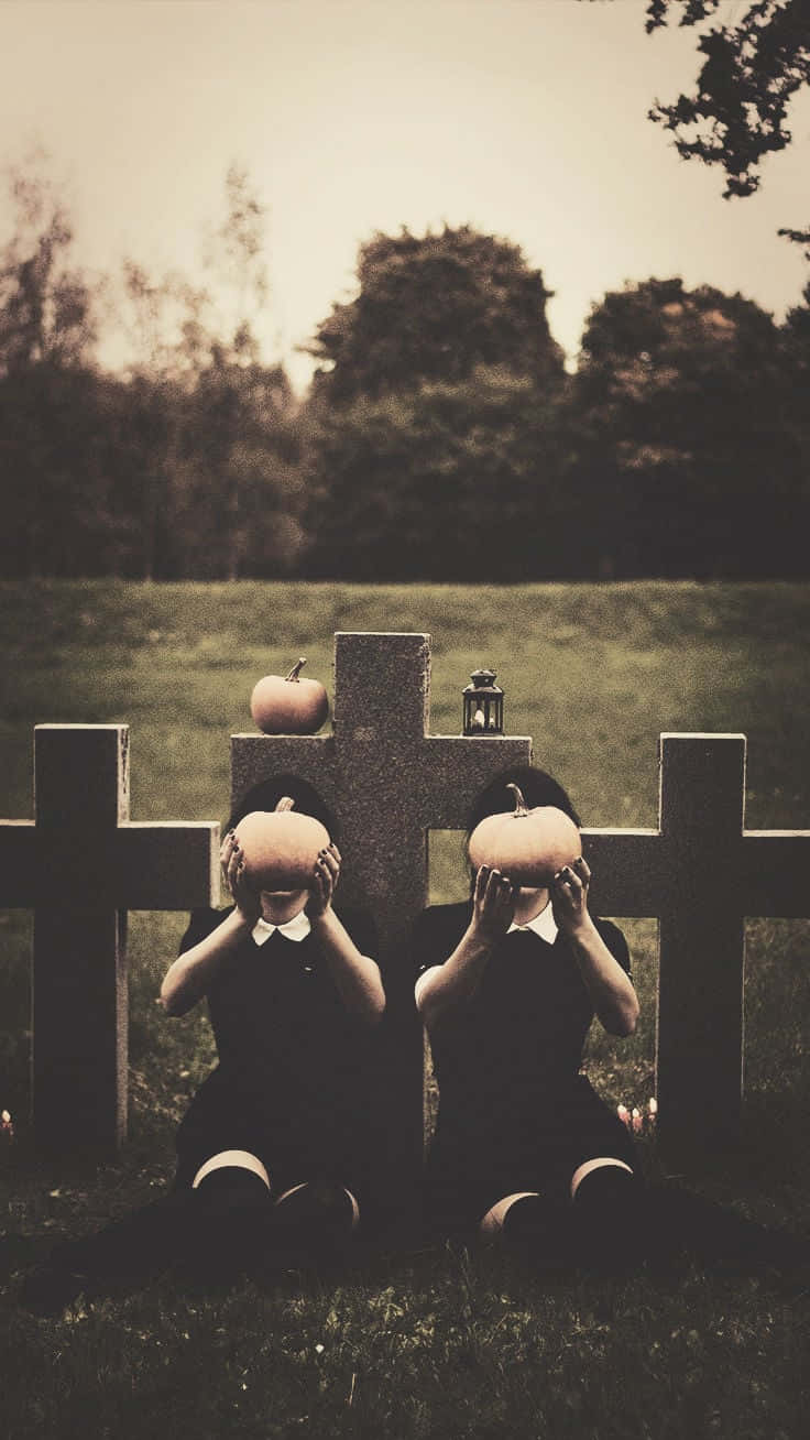Spooky iPhone 桌布 壁紙 — 兩個穿著黑色連衣裙的孩子坐在草地上，將南瓜舉在臉前，旁邊有石頭十字架，上面有南瓜和燈籠。
