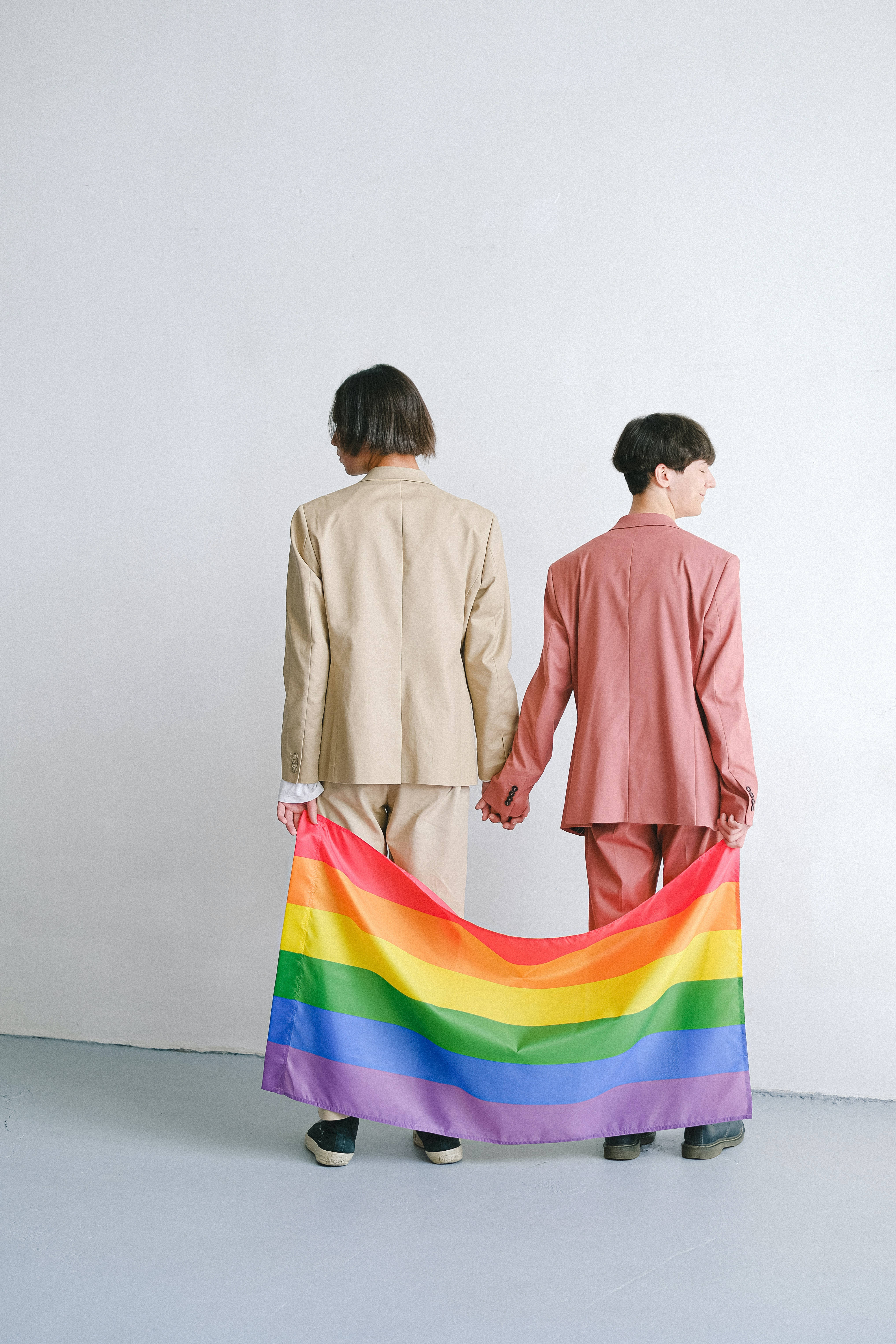Lesbian Flag 桌布 壁紙 — 兩個穿西裝的人手牽手，並在純白牆前舉起彩虹旗。