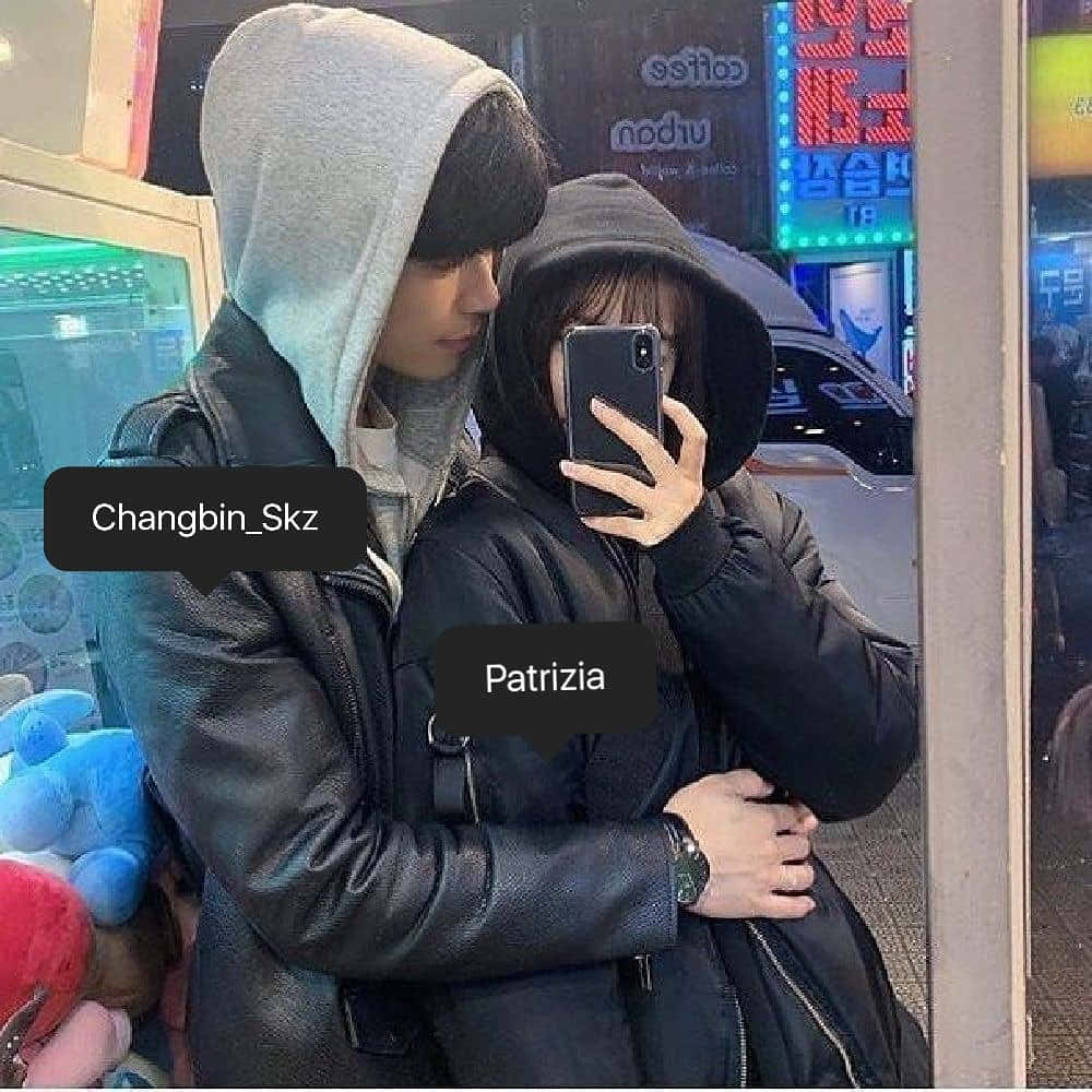 Matching Couple Profile 桌布 壁紙 — 兩個穿連帽衫的人在夜晚霓虹燈照亮的城市街道上自拍，附近有發光的招牌和絨毛玩具。