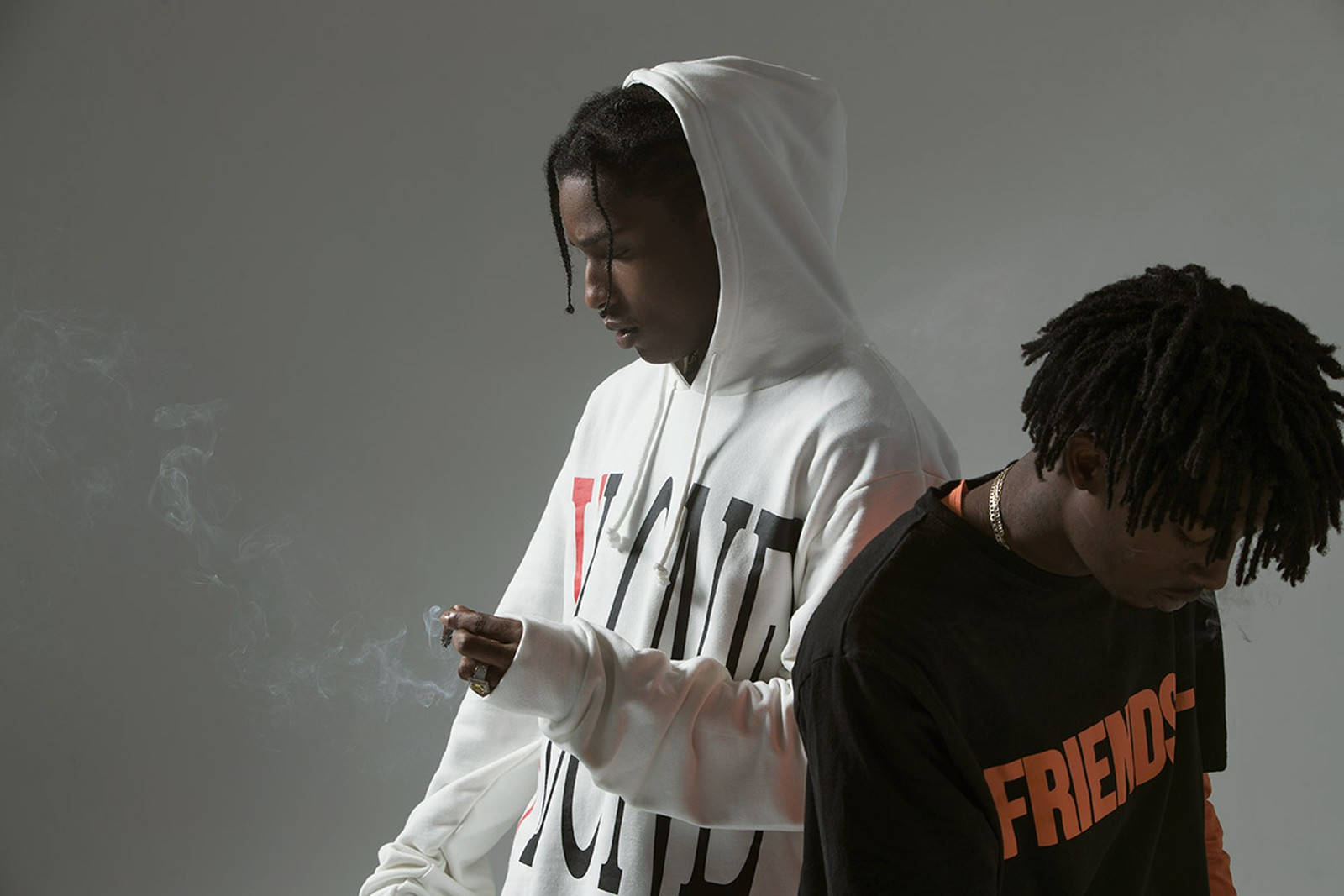 Vlone 桌布 壁紙 — 兩個穿連帽衫的年輕男子靠著灰色牆壁站立，一人抽煙，另一人低頭，兩人均表情嚴肅。
