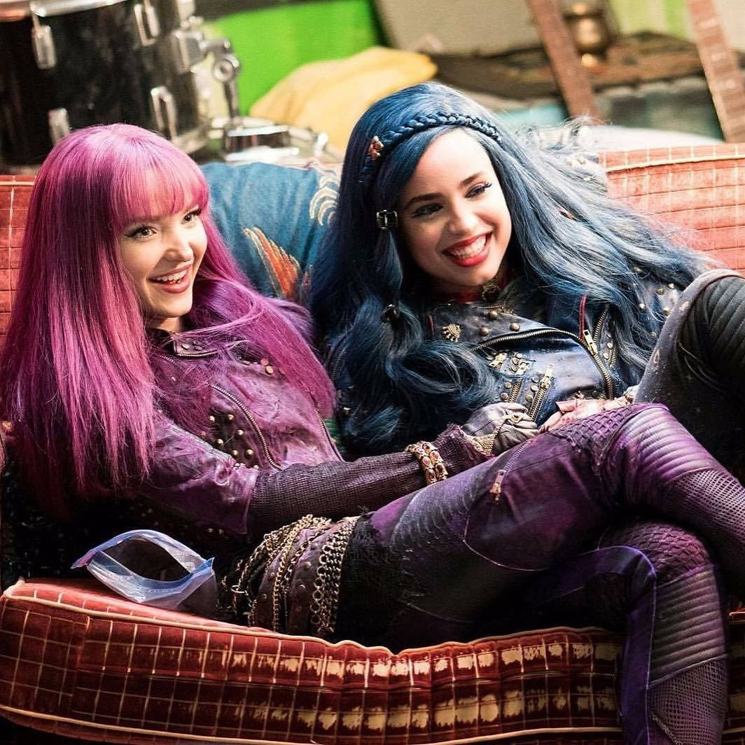 Descendants 桌布 壁紙 — 兩個笑容滿面的女孩，一頭鮮豔的粉紅色和藍色頭髮，在色彩繽紛的房間裡懶洋洋地躺在圖案沙發上。