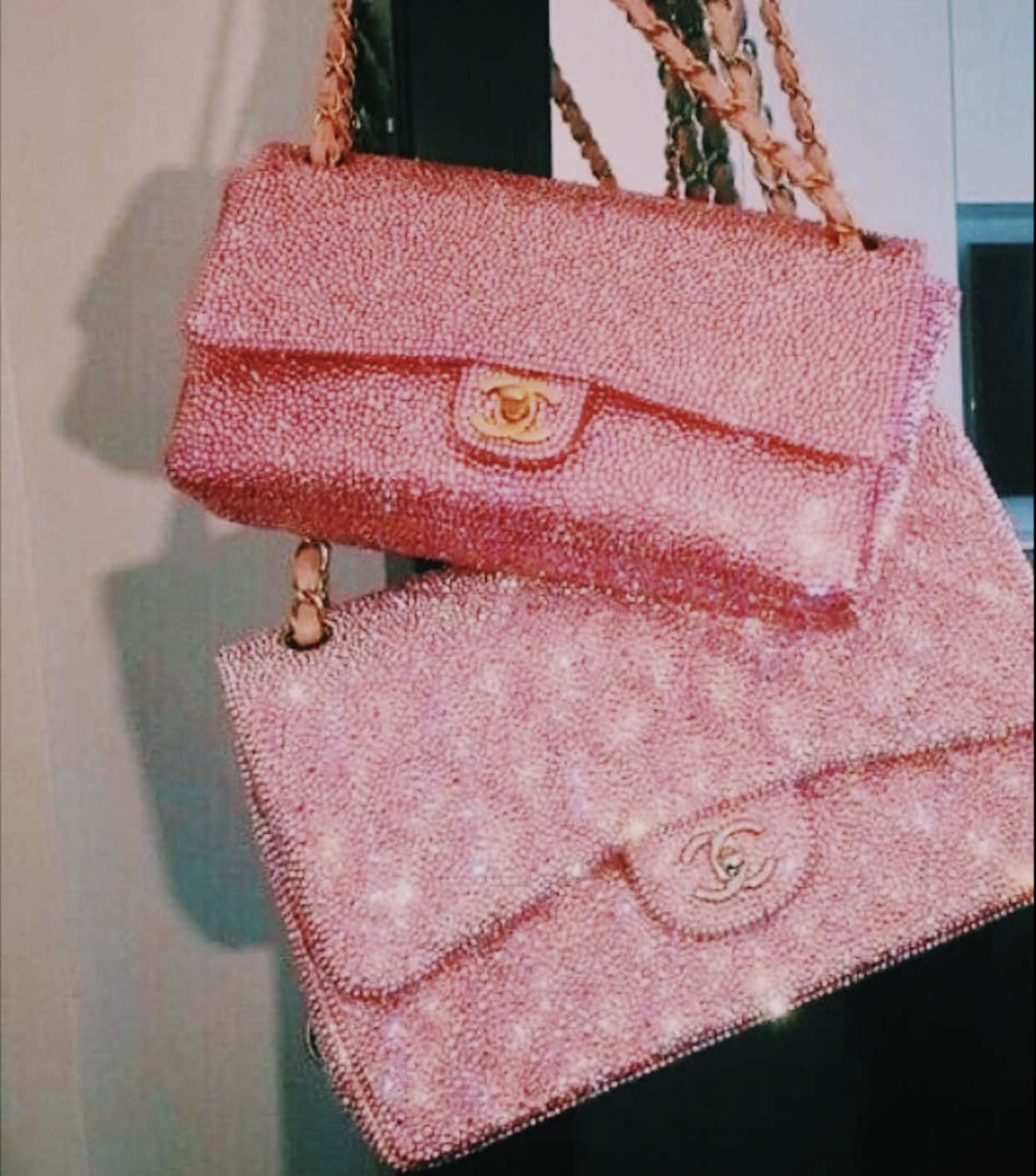 閃爍 桌布 壁紙 — 兩個粉紅色珠飾 Chanel 手袋，搭配金色金屬配件，掛在素色牆上，在柔和燈光下閃閃發光。