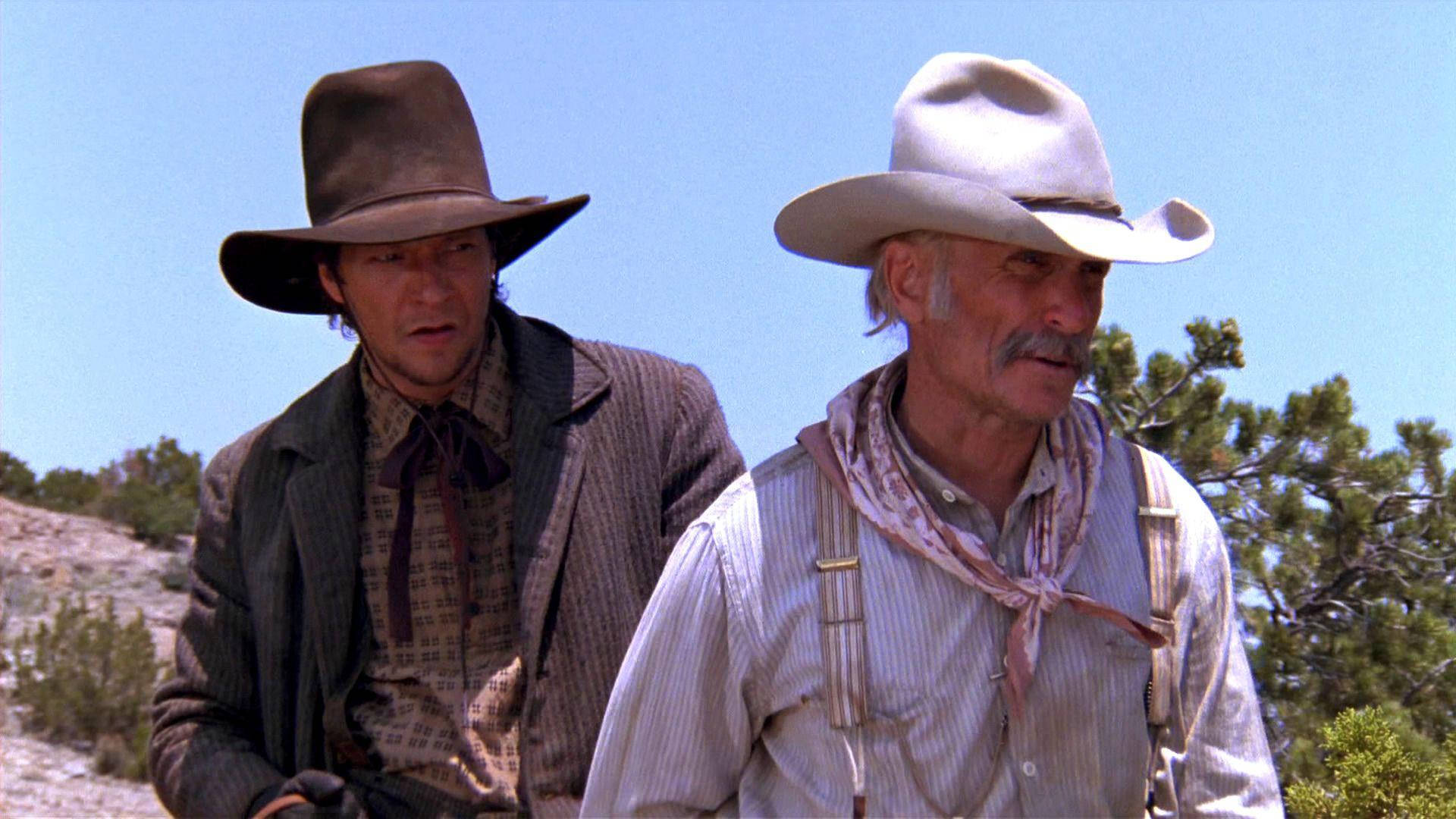 Cowboy 桌布 壁紙 — 兩個粗獷的牛仔戴著塵土飛揚的帽子，並肩站在明亮的藍天下，稀疏的綠樹和岩石地形。