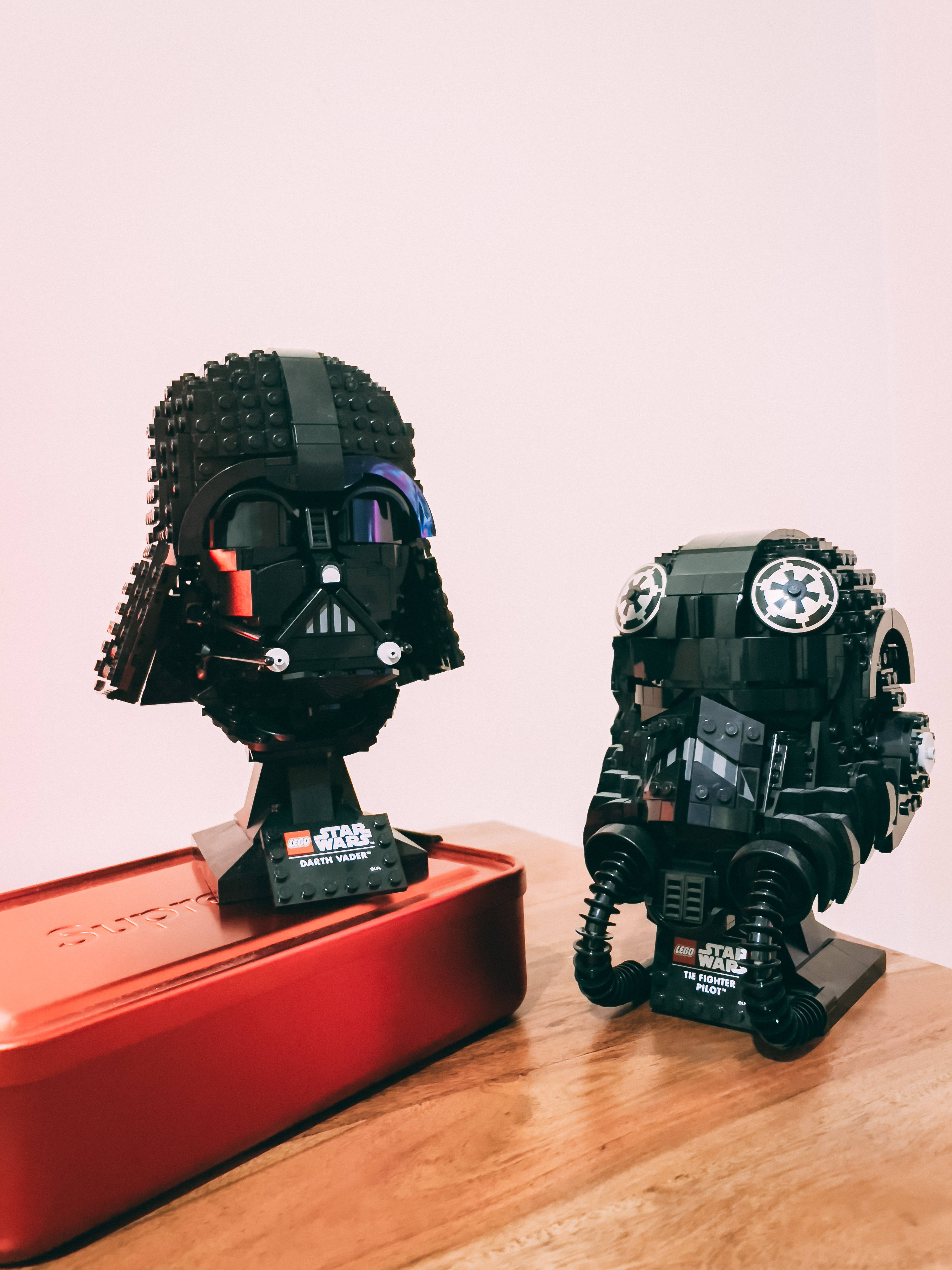 星際大戰 桌布 壁紙 — 兩個精細的LEGO星際大戰頭盔達斯維達和TIE戰鬥機飛行員放在木桌上，旁邊有一個紅色盒子，背景是淺色牆壁。