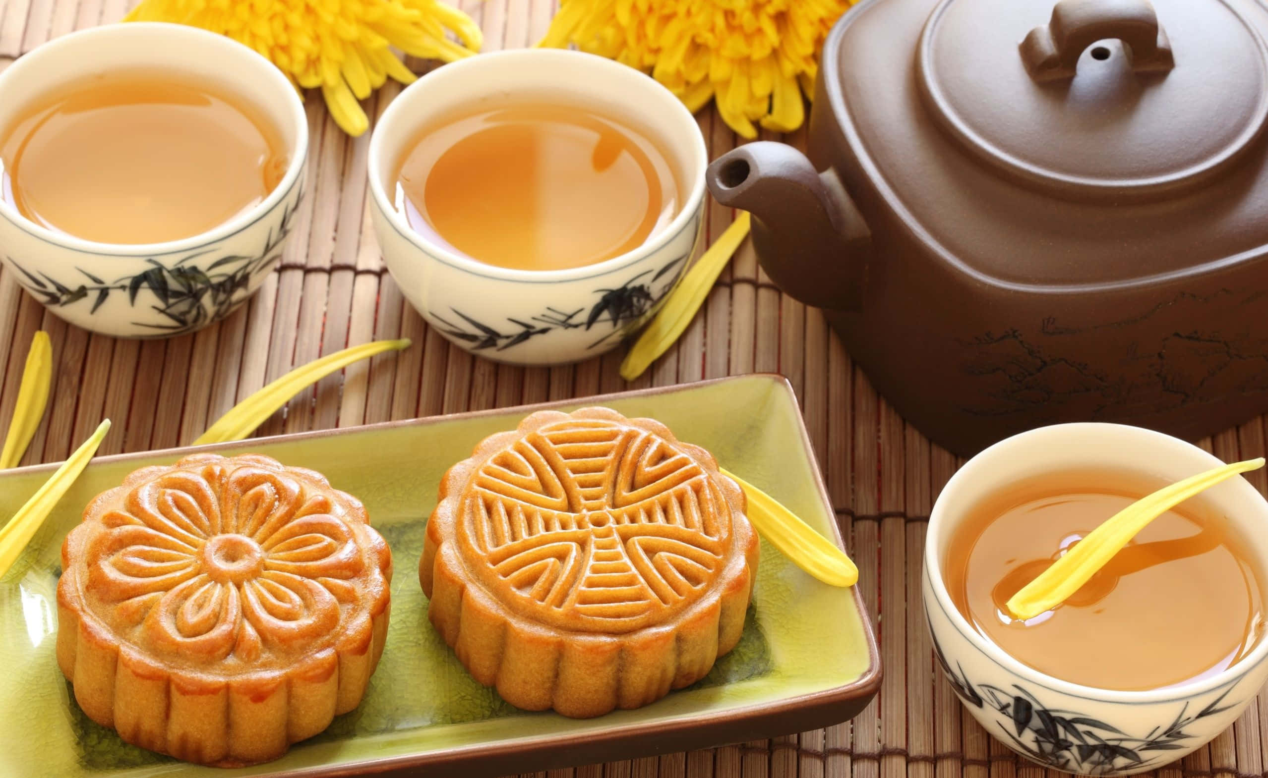 tea 桌布 壁紙 — 兩個精緻圖案的月餅放在綠色盤子上，旁邊有茶杯和陶壺，周圍是竹蓆上鮮豔的黃色菊花。