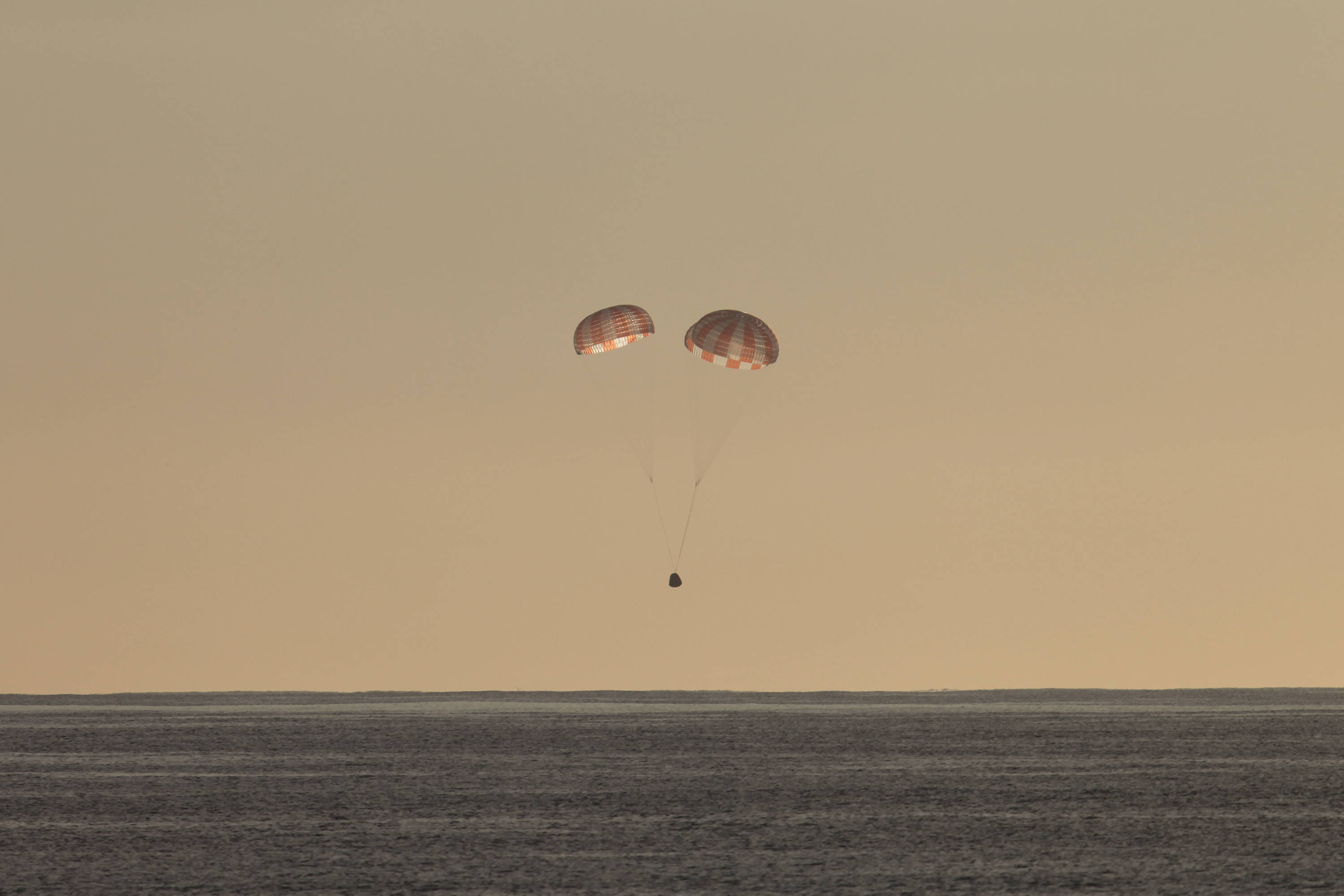 Aerospace 桌布 壁紙 — 兩個紅白條紋降落傘在朦朧的橙色天空下，緩慢降落在廣闊平坦的沙漠上。